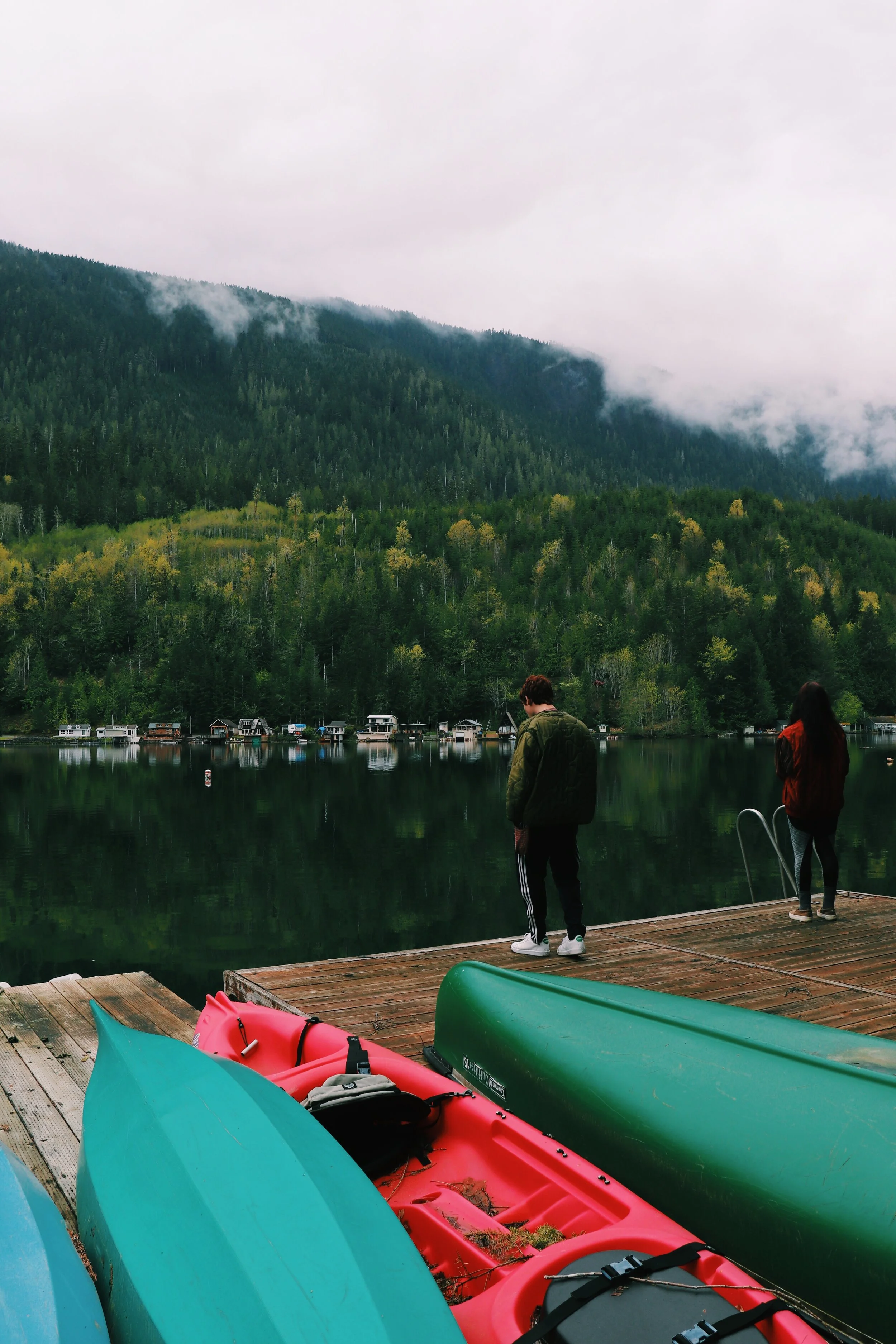 People standing on a wooden dock by a lake with colorful kayaks in the foreground, surrounded by lush green forested mountains under a cloudy sky. North West Washington State
