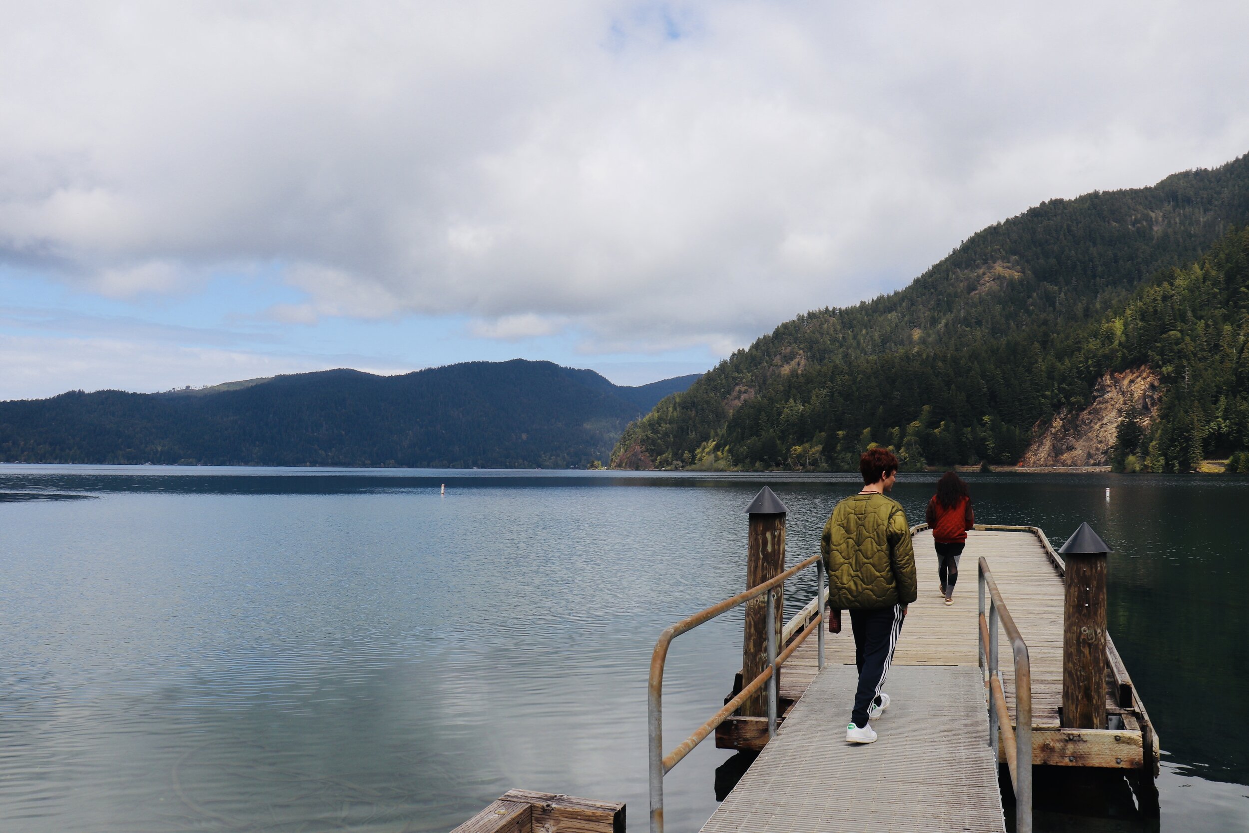 Two people walking on a wooden dock over a calm lake with forested mountains in the background.