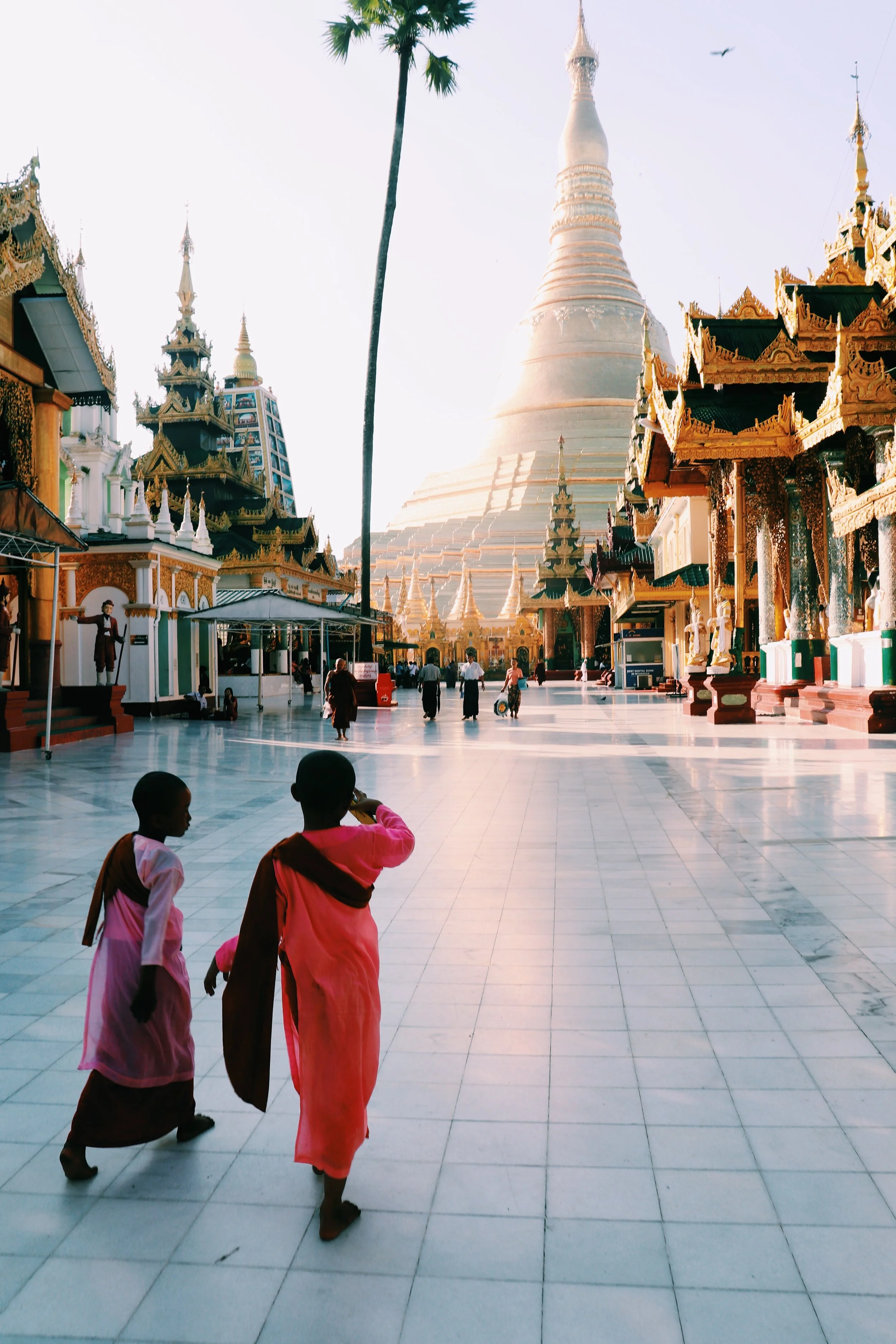 Young boys walking in front of a large traditional Buddhist temple with gold accents, multiple spires, and a large stupa in the background. Yangon Myanmar 