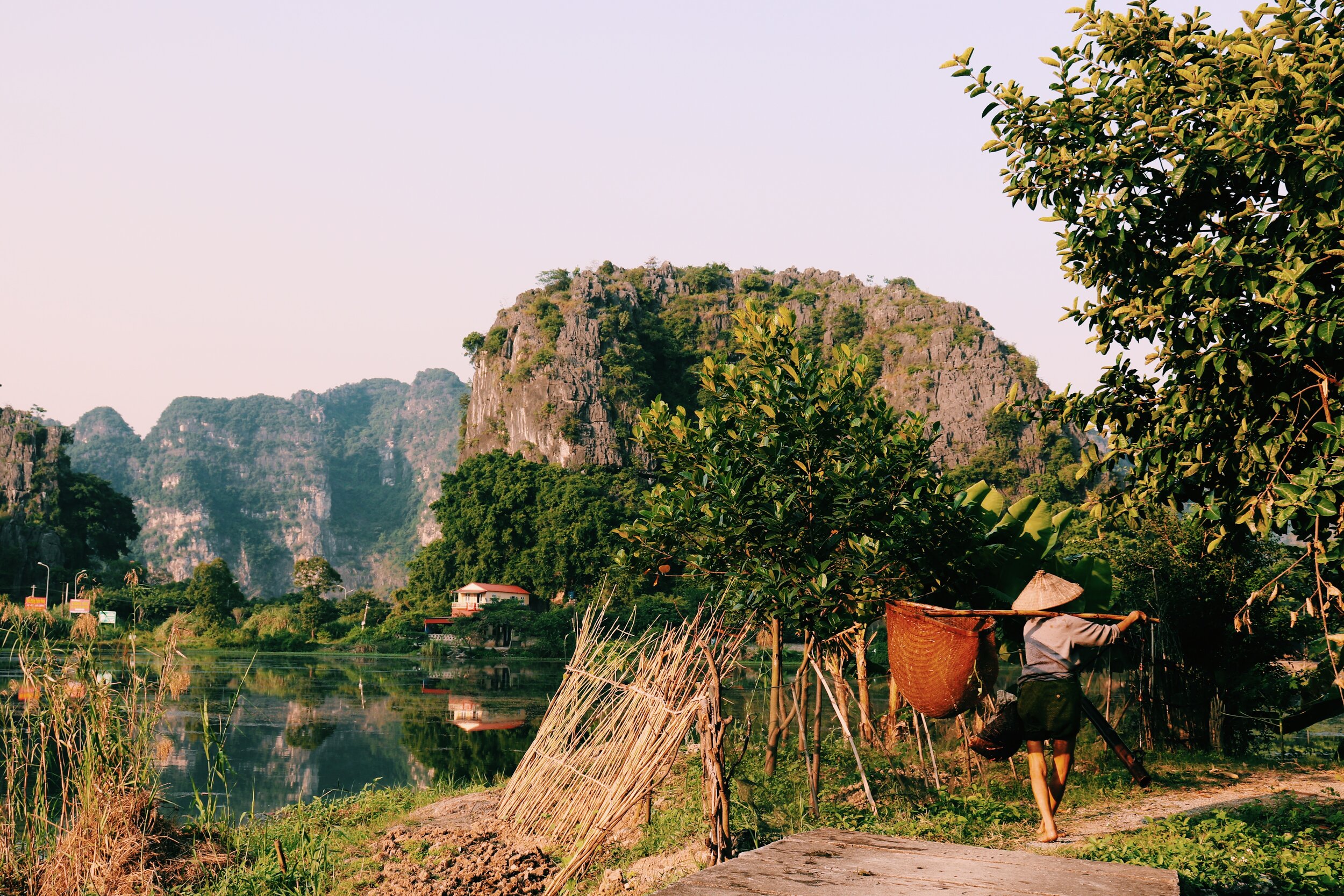 A person carrying a pole and basket walks along a rural path next to a body of water. The scene is set against lush greenery, tall trees, and towering limestone karst mountains in the background, with a partly cloudy sky overhead.