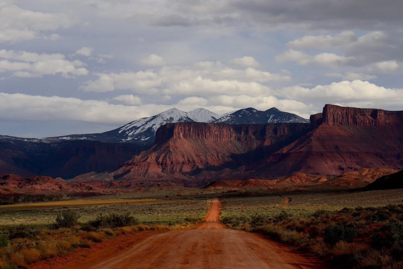 Dirt road leading towards rugged red rock formations and snow-capped mountains under cloudy sky. Moab Utah