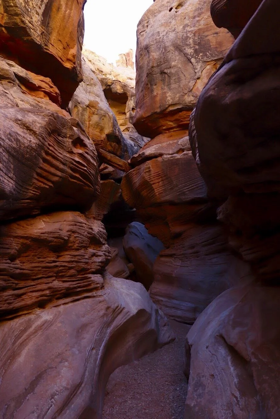 A narrow slot canyon with layered sandstone walls in shades of orange, red, and tan, with a small sandy path running through the center. Moab Utah