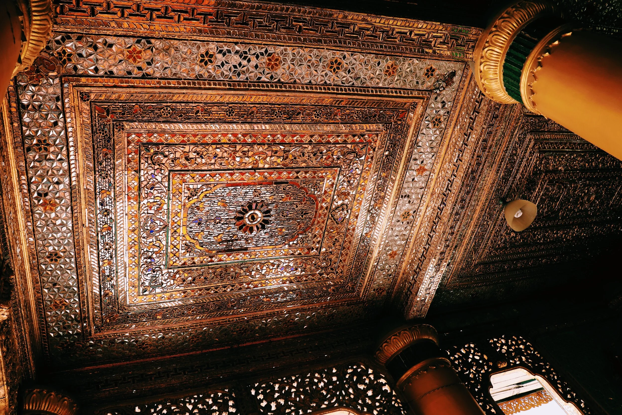 Intricate, gold-embellished ceiling with geometric and floral patterns, featuring columns with ornate capitals and detailed carvings, in a traditionally decorated interior. Yangon Myanmar