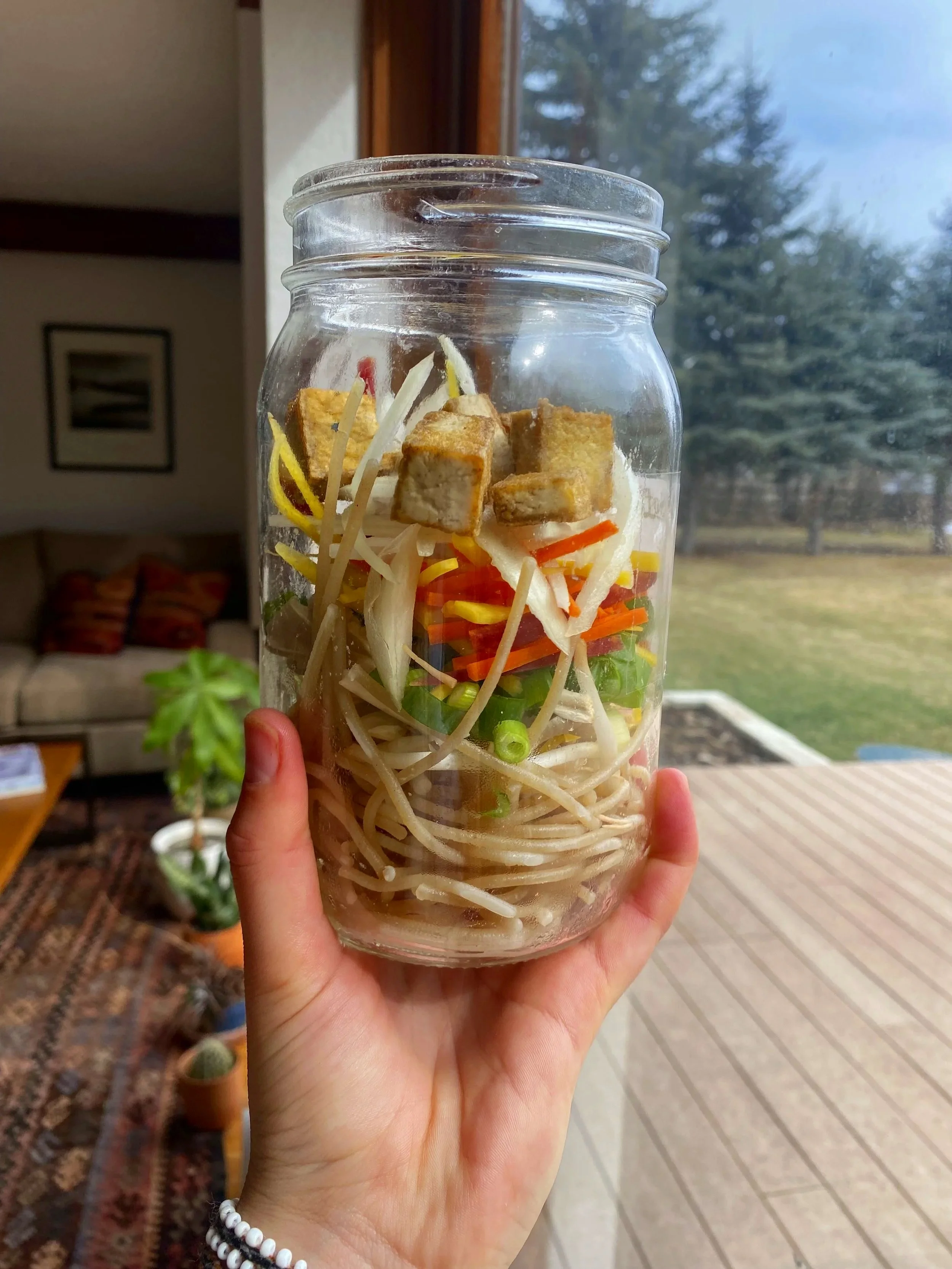 Private Chef in Aspen Colorado Roaring fork valley holding a glass jar filled with layered ingredients including noodles, chopped vegetables, and tofu, with a scenic outdoor background.