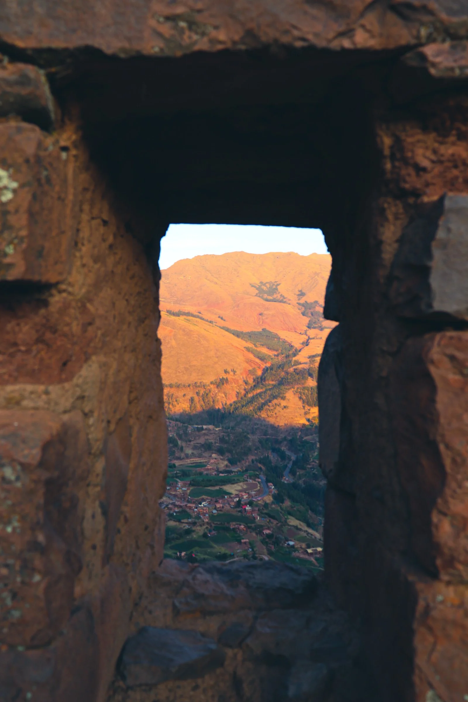 View of a mountain landscape through a small rectangular stone window, showing rolling hills with patches of green, reddish-brown terrain, and a small village at the base. Pisac Peru