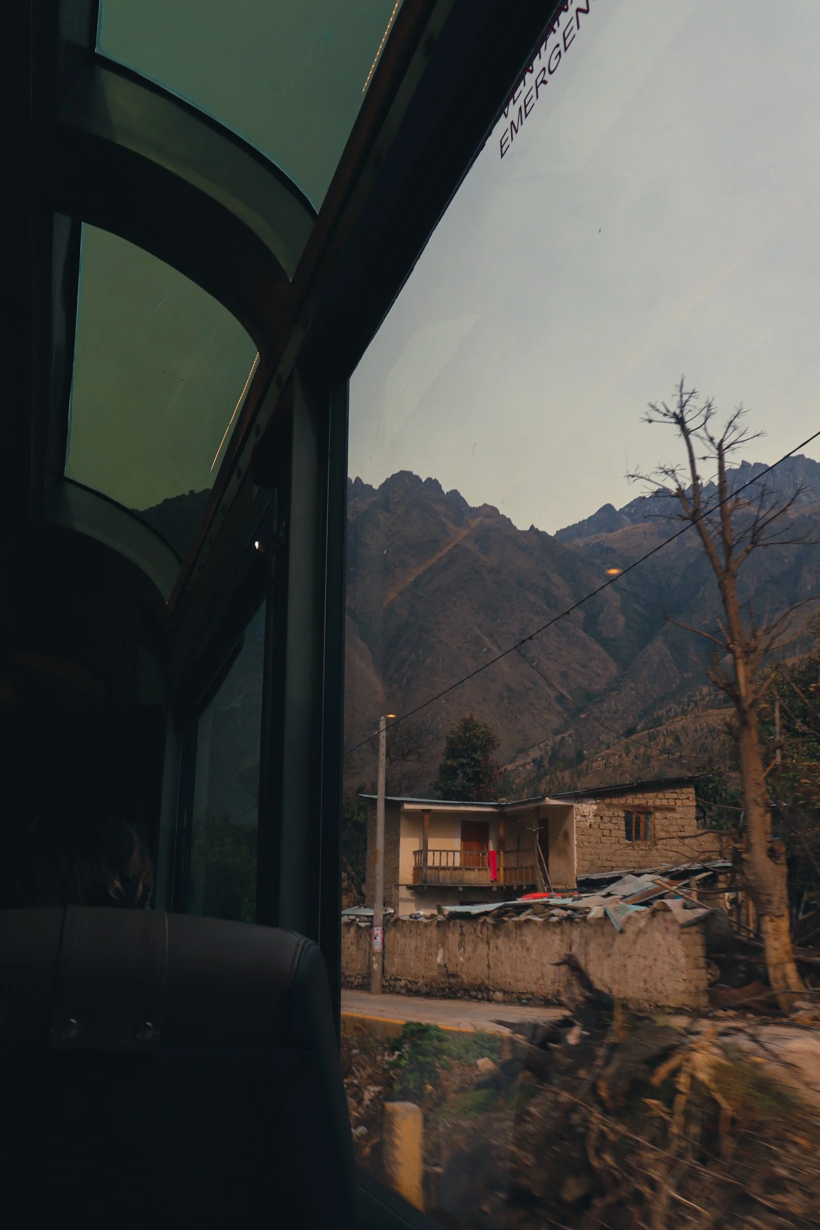 A view from inside a bus window showing a mountainous landscape with rocky peaks, a small house, and a tree with bare branches. Peru