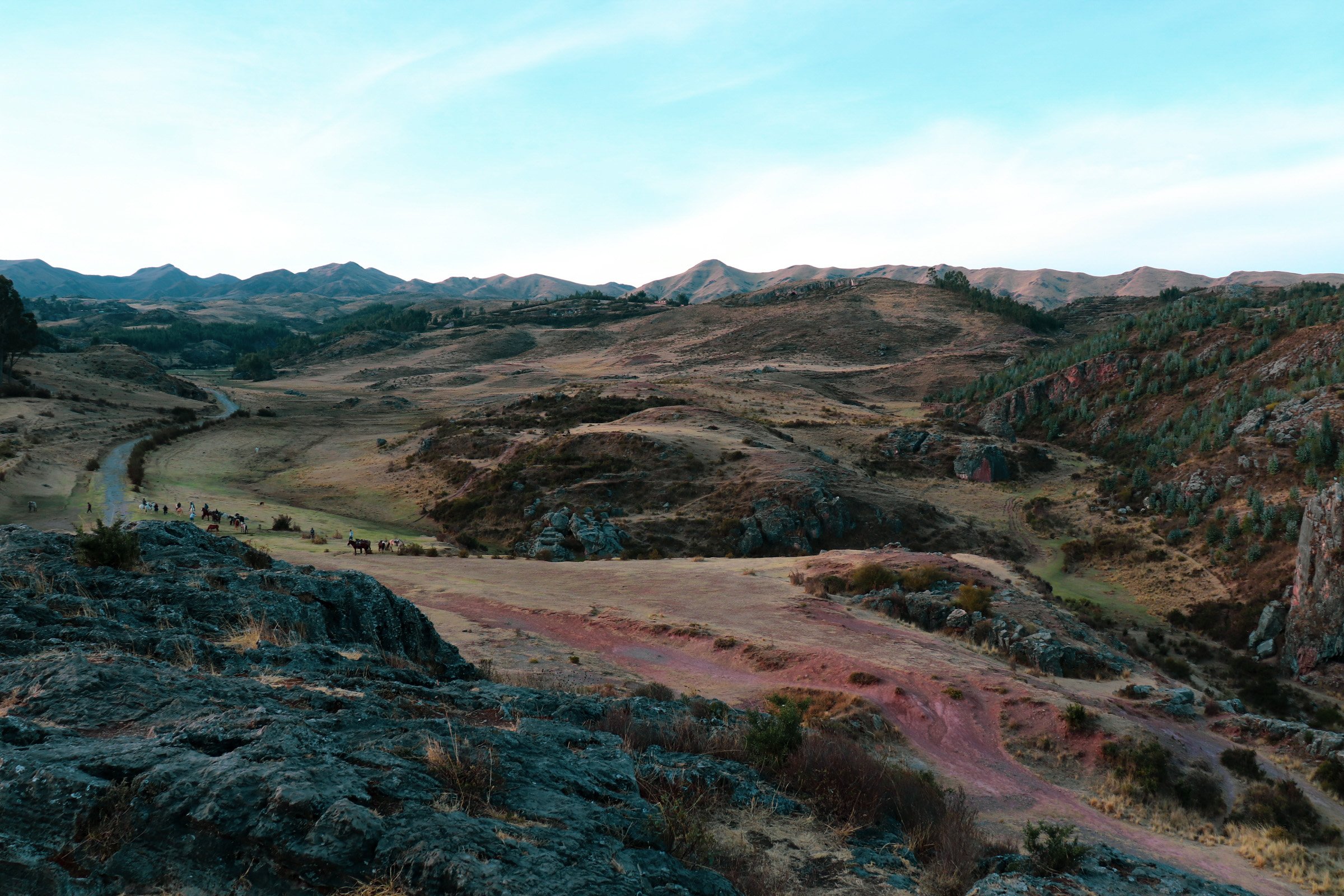 A scenic landscape of mountains and rolling hills with a dirt path winding through the terrain. There are horses visible in the distance and sparse vegetation across the area. Cusco Peru