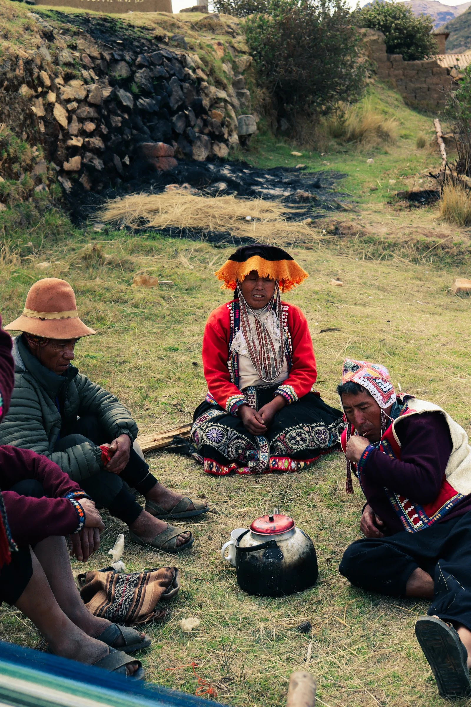 Group of indigenous people sitting on the ground outdoors, wearing traditional clothing and hats, with a kettle in the center, near a stone wall and paths. Pisac Peru