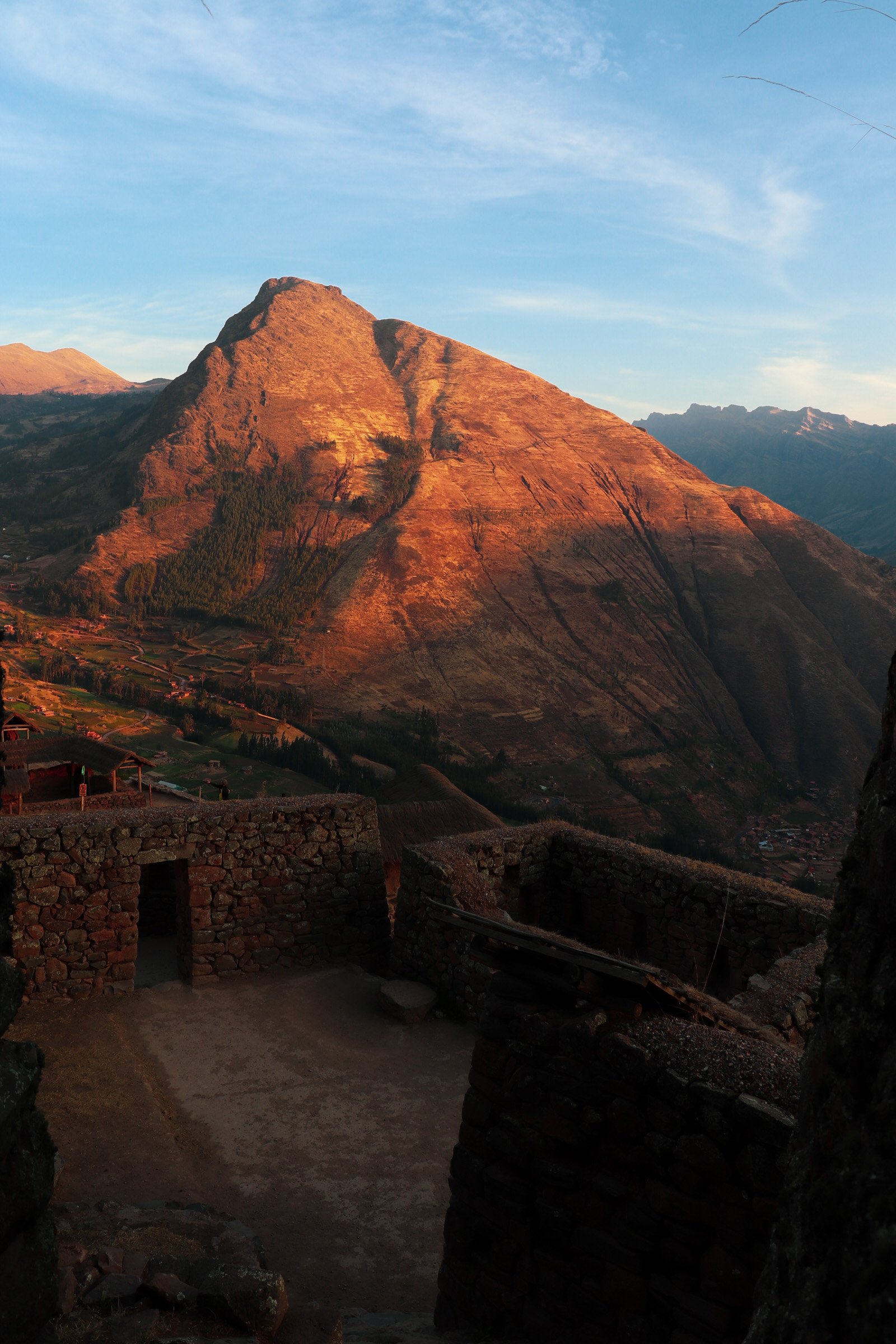 A mountain scene during sunset with a large mountain illuminated in orange hues, and ancient stone ruins in the foreground. Pisac Peru