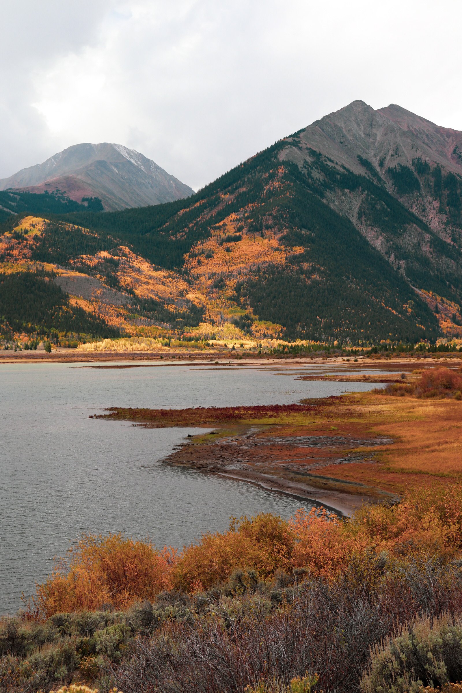 Scenic landscape of a lake with autumn-colored shrubbery in the foreground, mountains covered with forests and patches of fall foliage, and a cloudy sky above. Twin Lakes Colorado