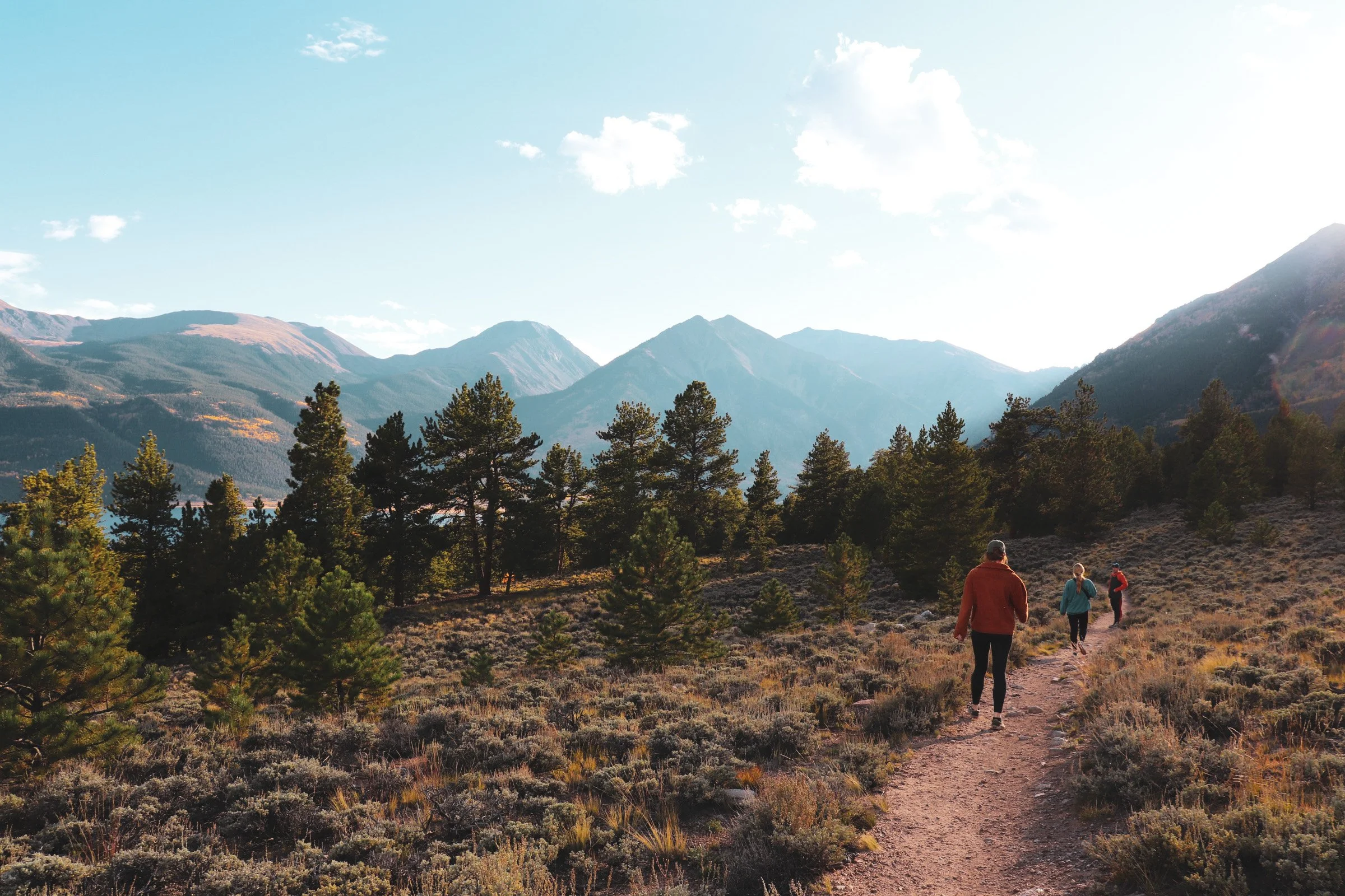 Three people hiking on a trail surrounded by trees and mountains under a partly cloudy sky. Colorado