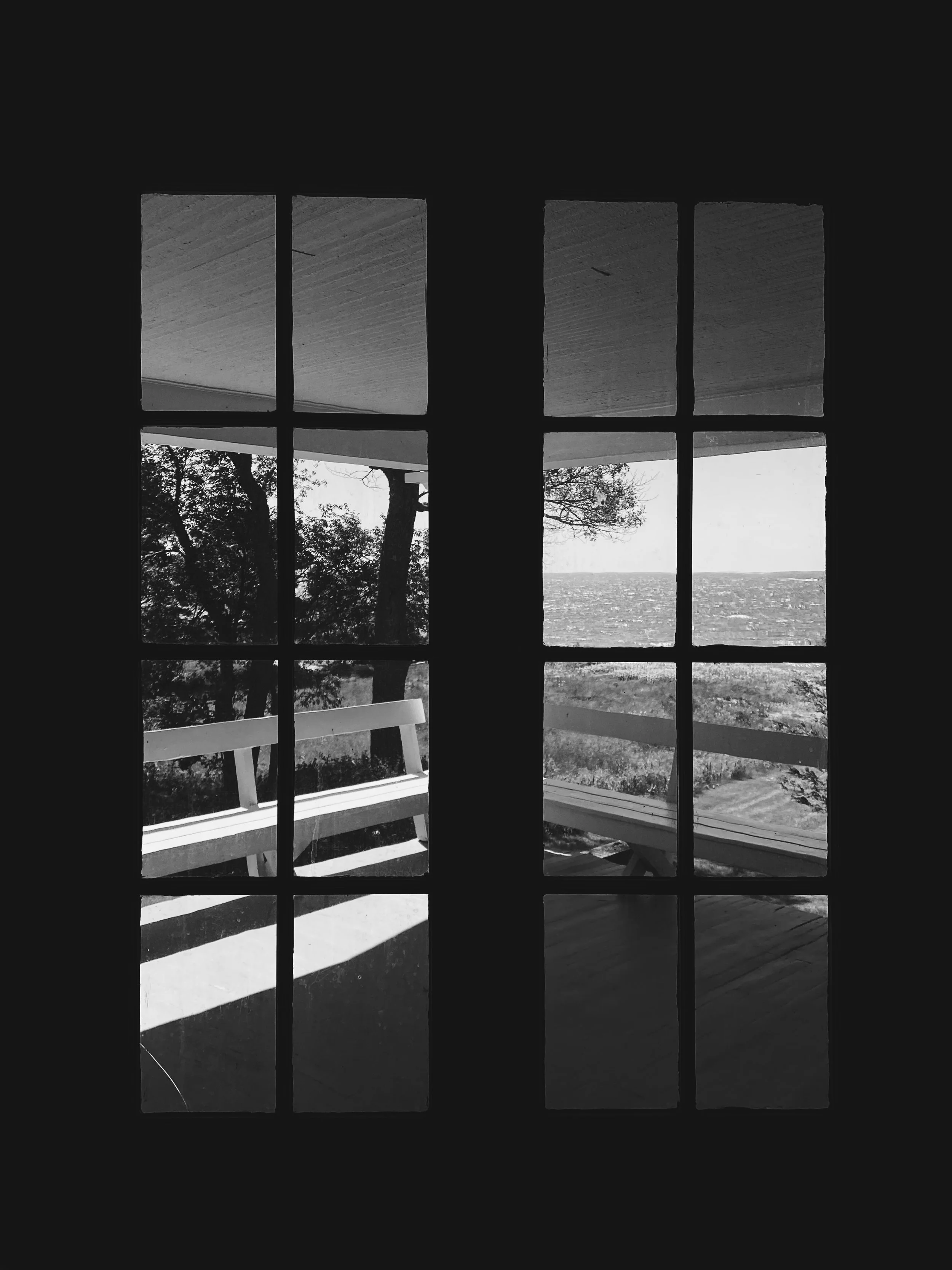 Black and white photo of a view through a multi-pane window showing trees, a boat, and water outside, with a bench on the porch outside.