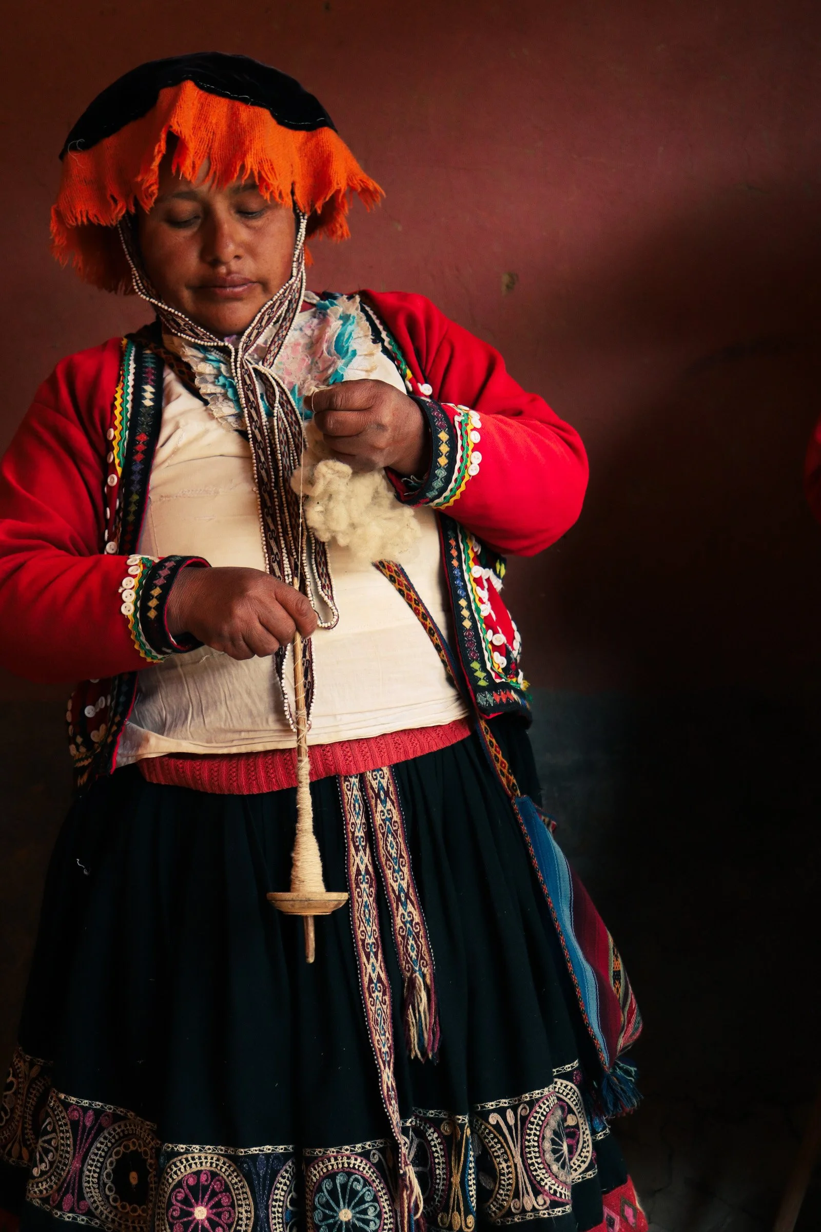 A woman wearing traditional indigenous clothing, including a colorful embroidered skirt, a red sweater, and an orange and black hat, is knitting with yarn.