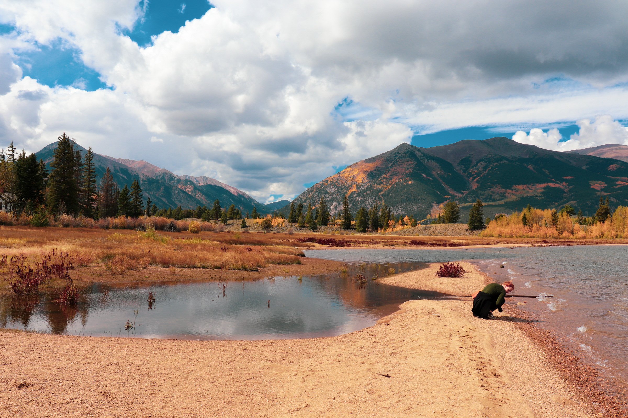 A person crouching on a sandy shore by a lake with mountains and pine trees in the background under a partly cloudy sky. Twin Lakes Colorado