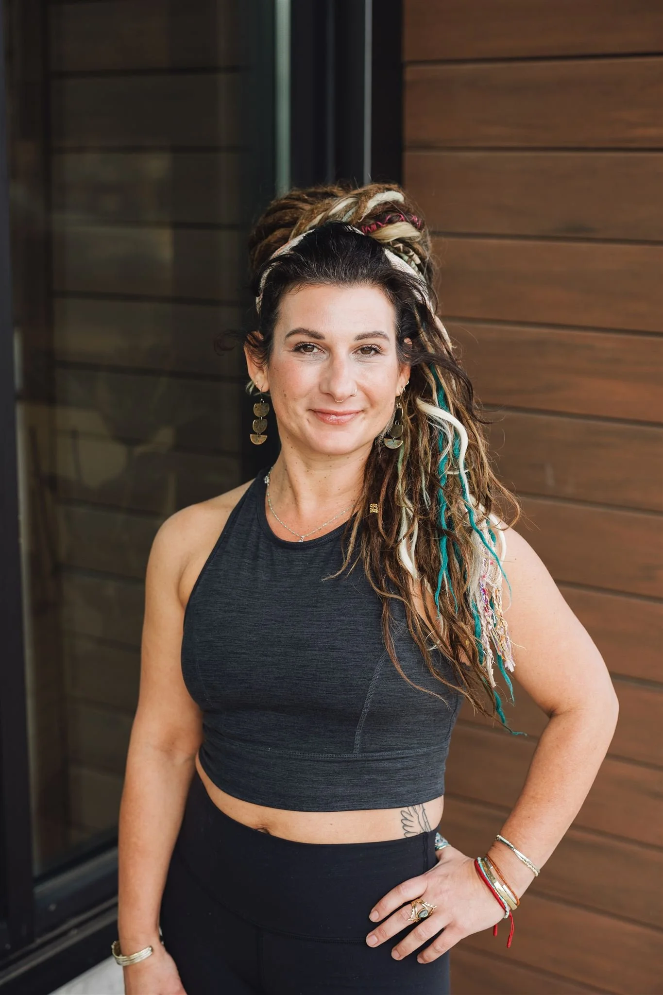 A woman with long, styled dreadlocks with colorful extensions, wearing a black athletic crop top and black pants, standing with her hand on her hip in front of a wooden wall.