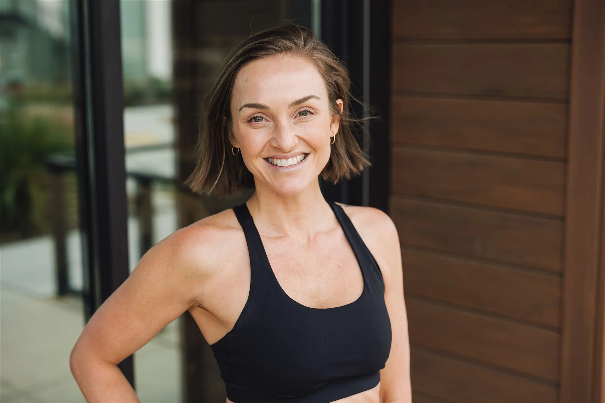 A woman with short brown hair, wearing a black sports bra and hoop earrings, smiling outdoors in front of a building with wooden siding.