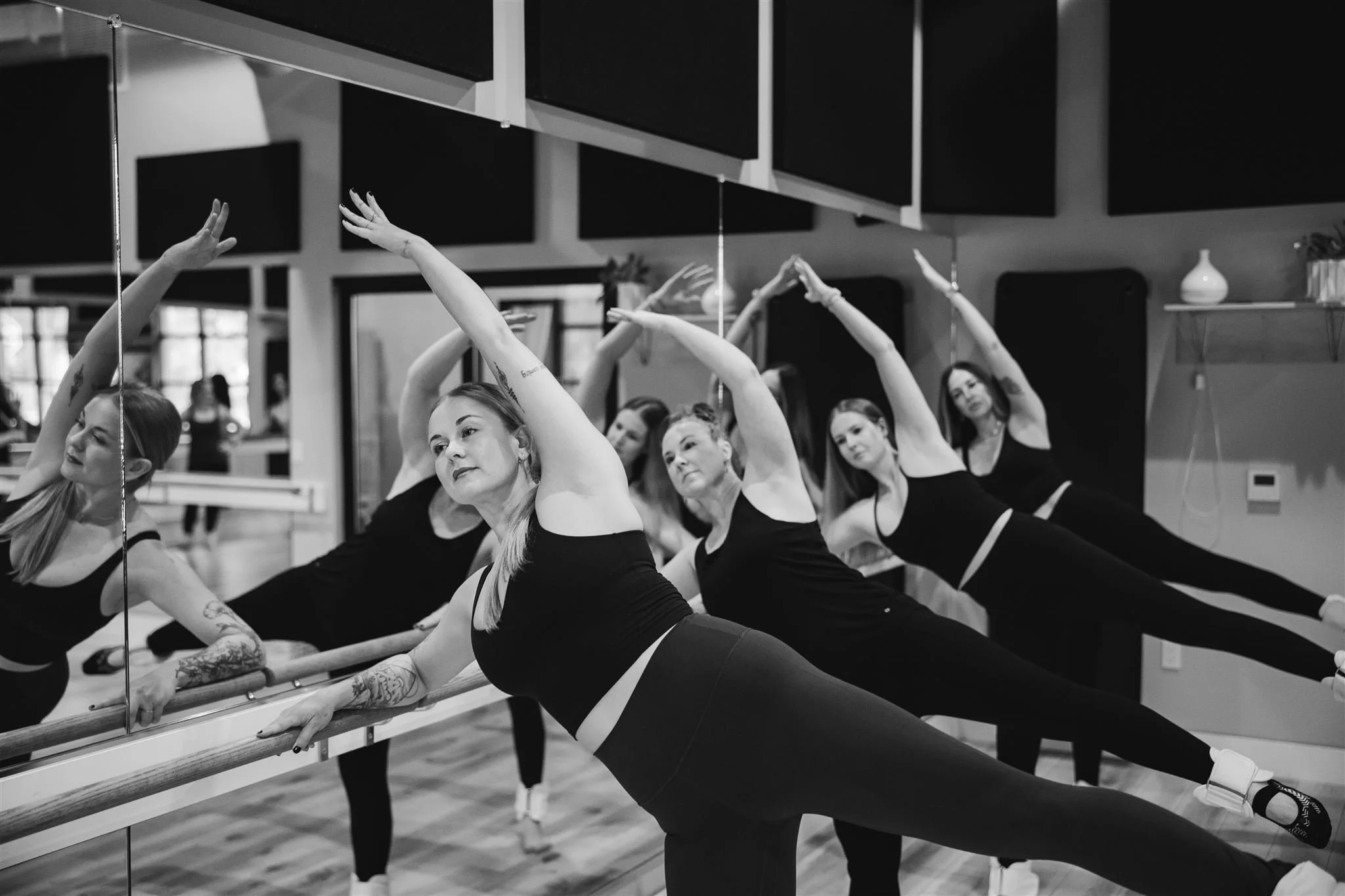 Group of women in black workout clothes stretching at a ballet barre in dance studio, practicing dance moves in front of mirror.