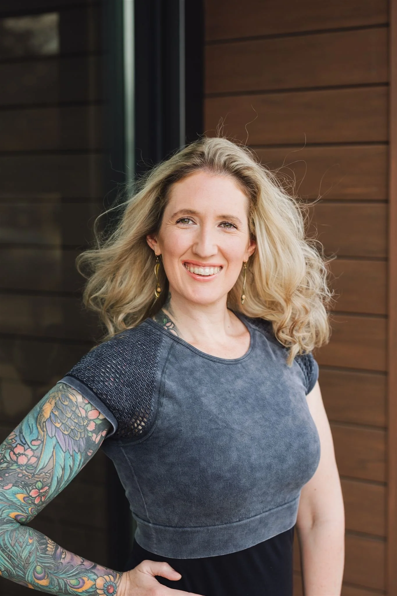 Smiling woman with blonde curly hair, wearing a gray athletic top with mesh shoulders and colorful tattoo sleeve, standing in front of wood panel and glass background.
