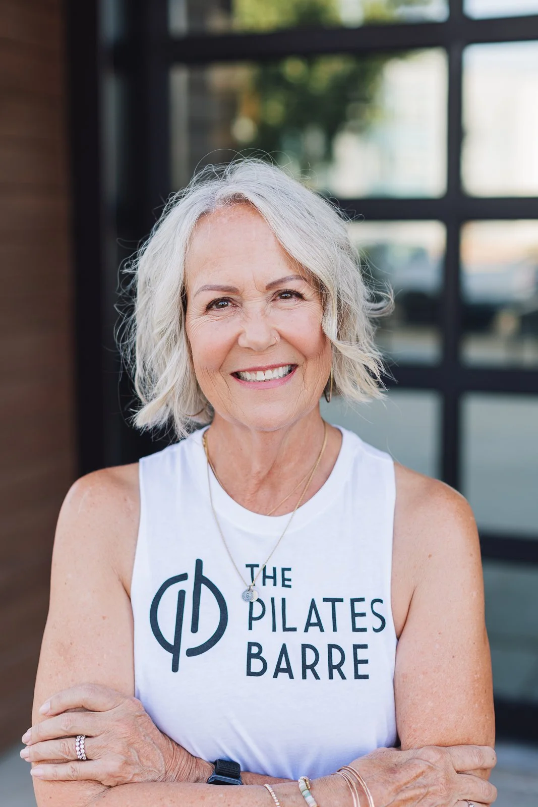 Smiling older woman in sleeveless shirt with text 'The Pilates Barre', standing outdoors near a building with glass windows.