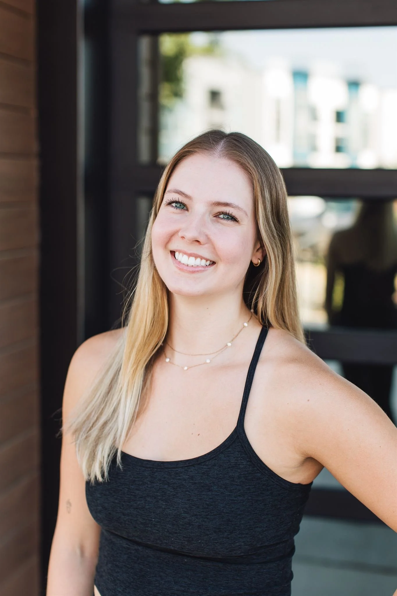 A young woman with long blonde hair and blue eyes smiling at the camera, wearing a black tank top and a layered necklace, standing outdoors in front of a glass door with a blurred cityscape background.