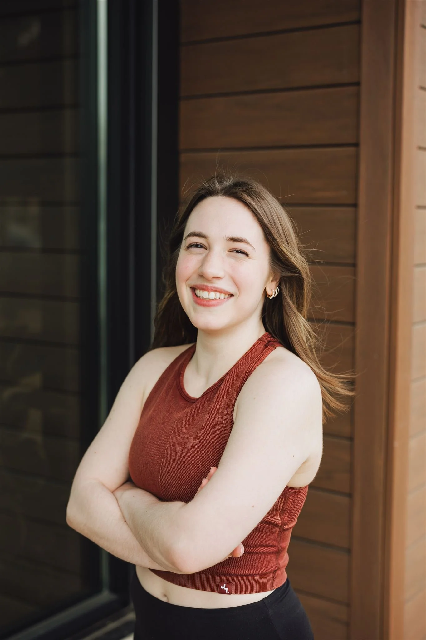 Young woman with long brown hair, smiling, wearing a rust-colored sleeveless top and black pants, standing outside near wooden siding.