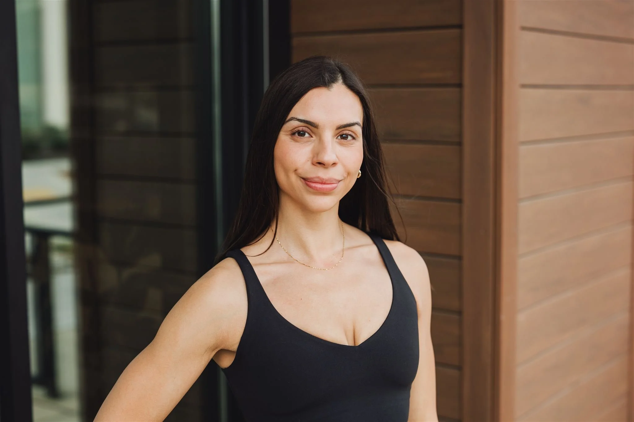 A woman with long dark hair, wearing a black sleeveless top and gold jewelry, stands outside near a wooden wall, smiling at the camera.