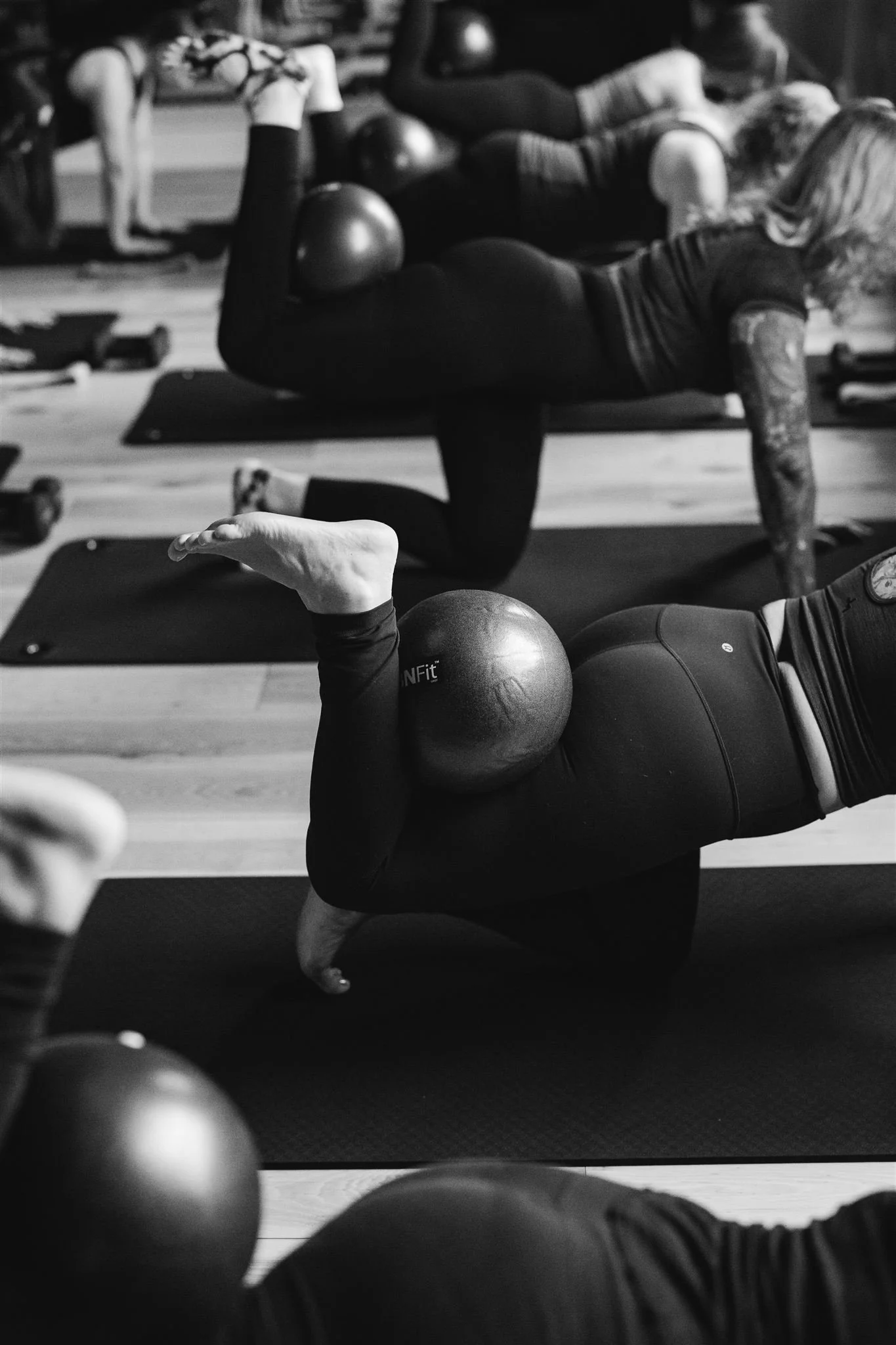 Women participating in a fitness class, lying on a mat and holding a ball on their back while performing a stretch or exercise.