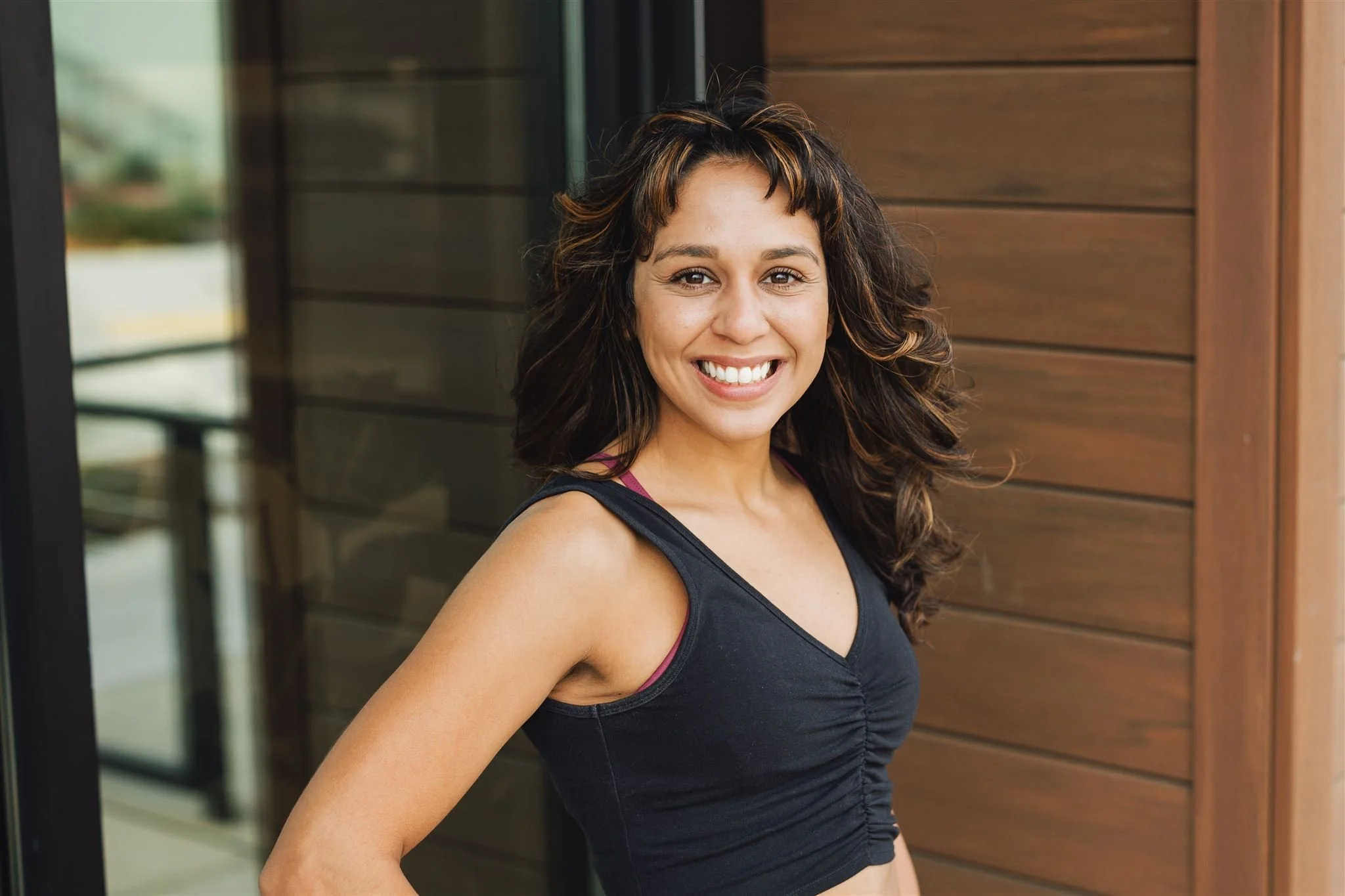 A smiling woman with wavy dark brown hair, wearing a black sports top, standing outside near a building with wooden paneling.
