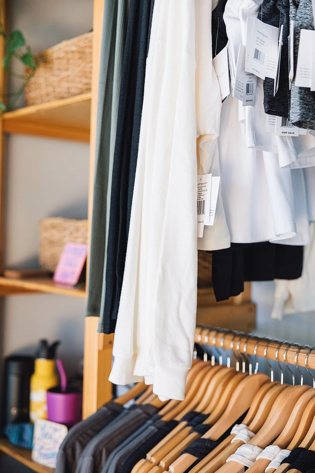 Clothing rack with white, black, and gray garments hanging on wooden hangers, with a wooden shelf in the background holding baskets and a pink sign.