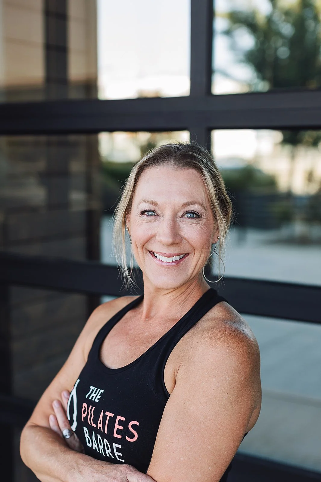 A smiling woman wearing a black tank top with the words 'The Pilates Barre' stands outdoors with her arms crossed in front of a modern building with large windows.