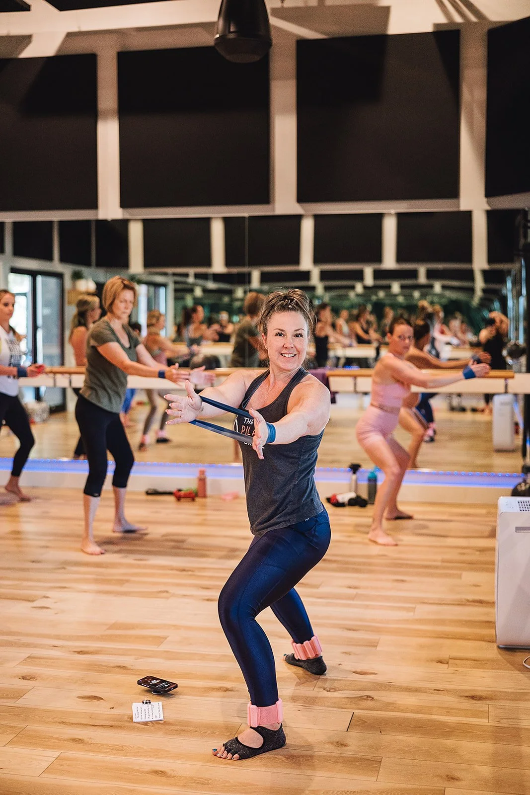 A group fitness class in a studio with women stretching and exercising, with a woman in the foreground smiling at the camera.