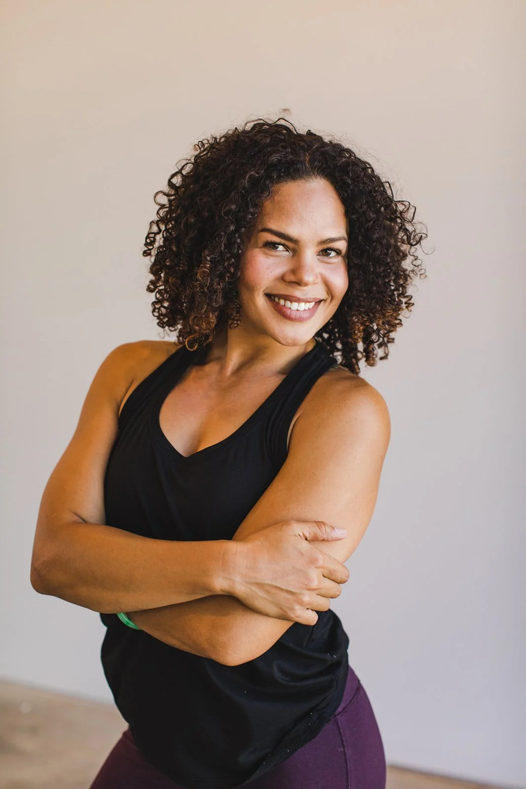 A woman with curly hair in a black athletic top and purple leggings smiling and crossing her arms.
