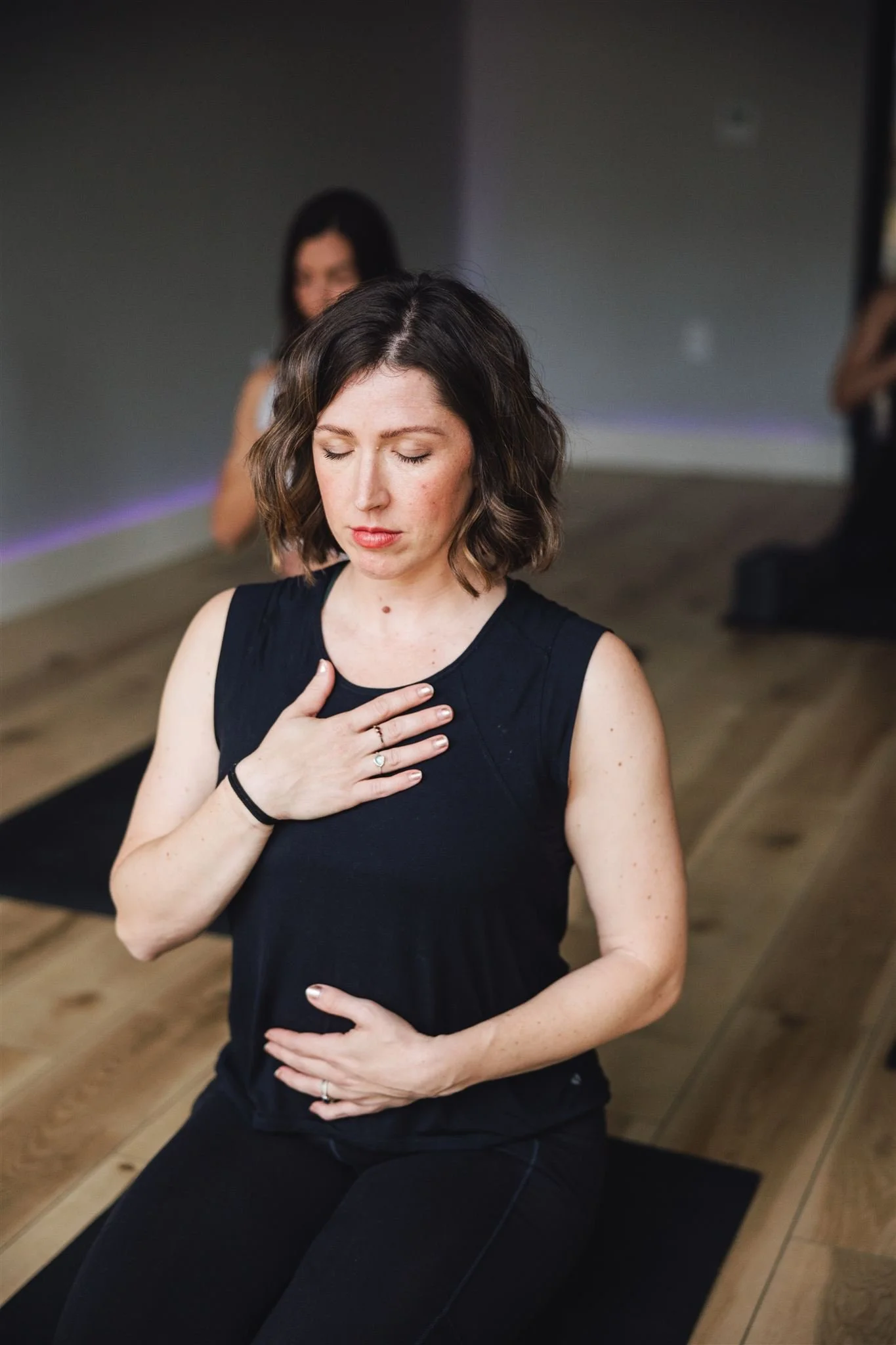 A woman practicing yoga, kneeling on a black mat with her eyes closed, one hand on her chest, and the other on her stomach, in a softly lit room.