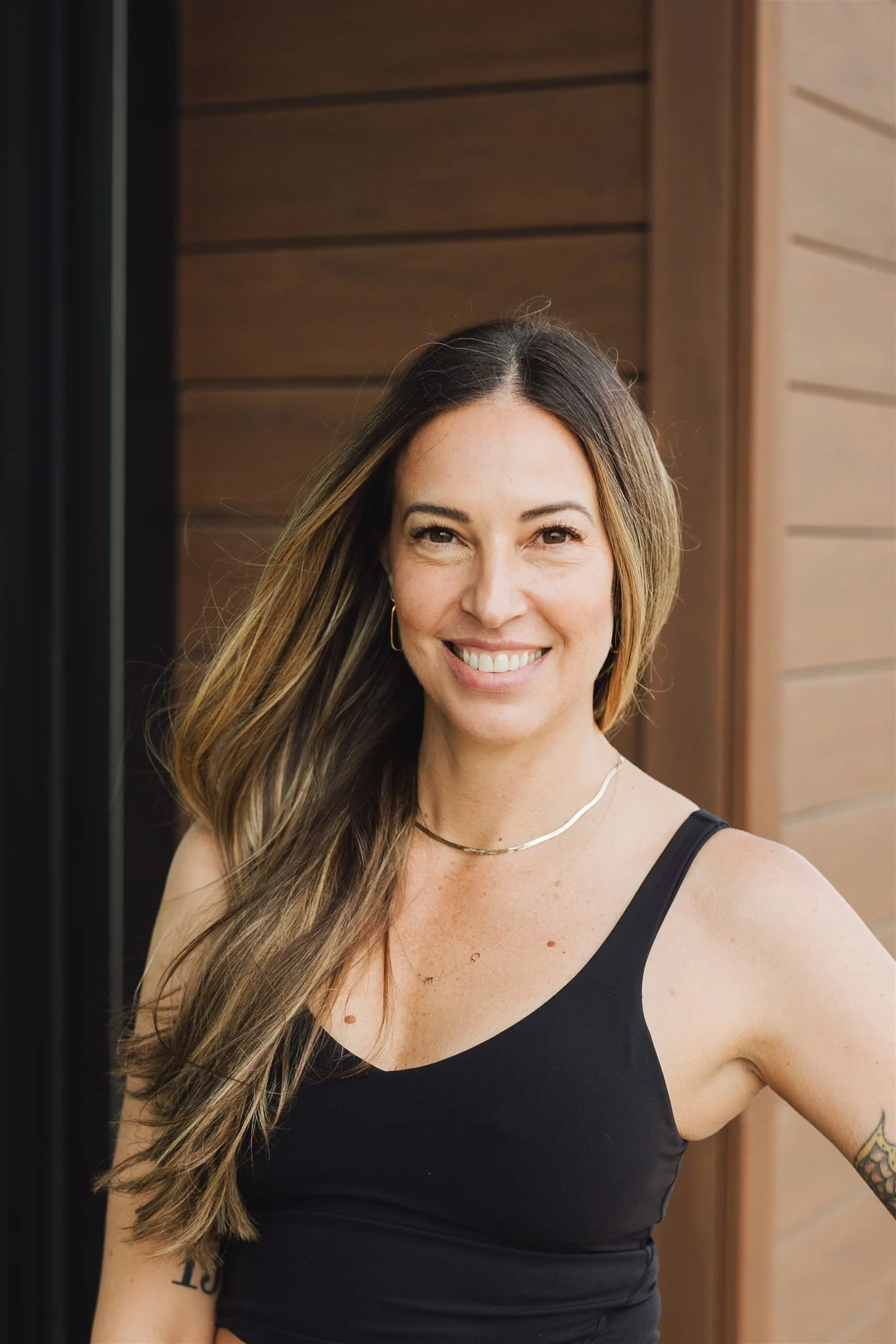 A smiling woman with long, brown hair wearing a black tank top and jewelry.