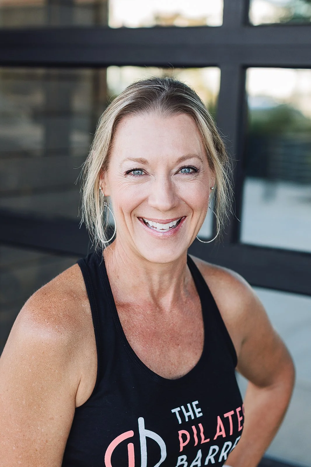 A smiling middle-aged woman with short blonde hair, wearing a black tank top with pink and white lettering, standing outdoors in front of a glass window.