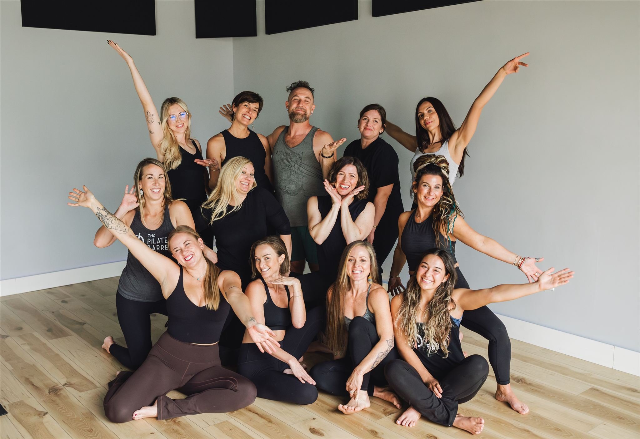 Group of energetic women and one man posing in a yoga or fitness studio, some kneeling and some standing, all smiling and celebrating, with a plain wall and wooden floor in the background.