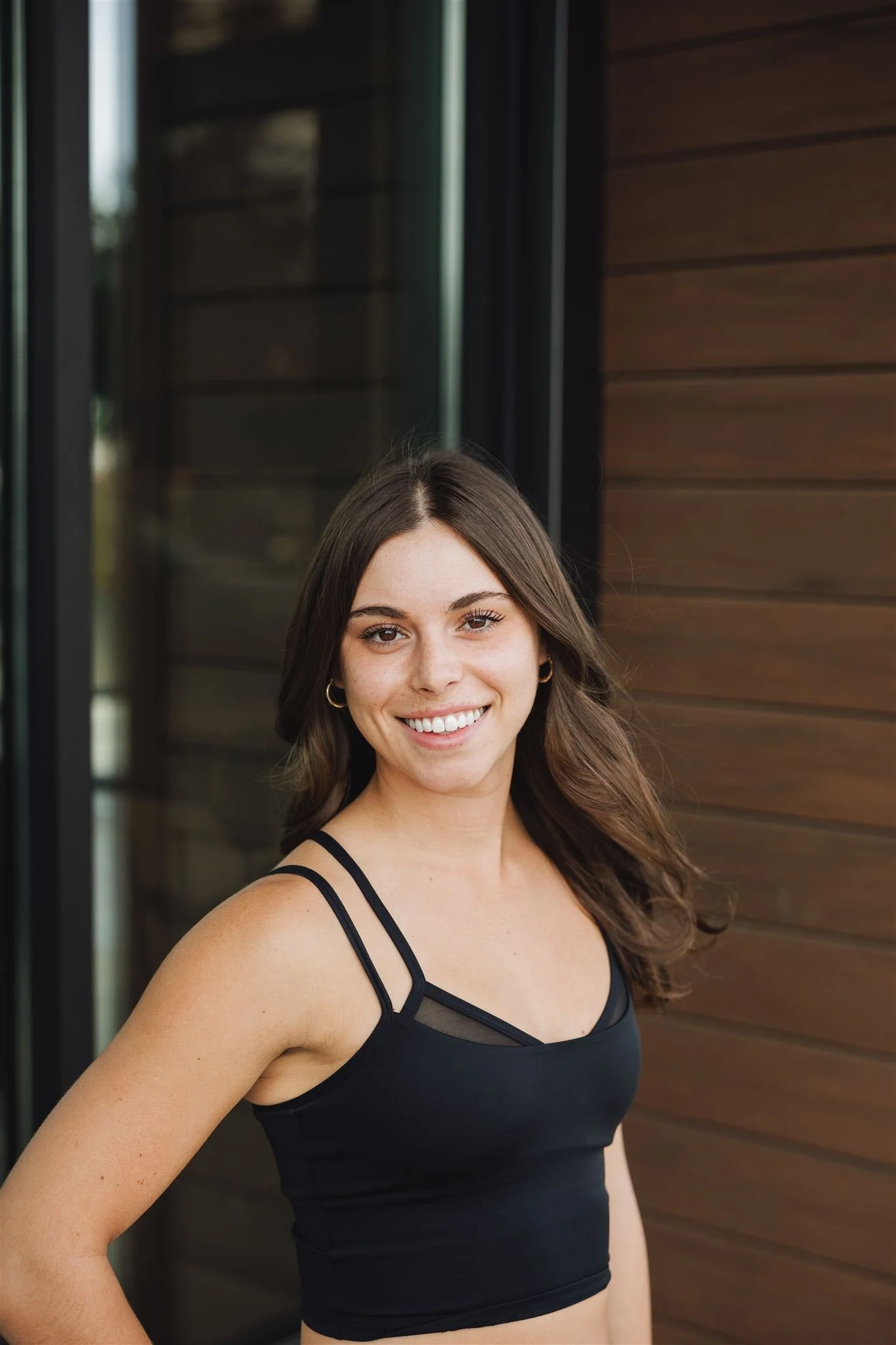 A woman with long brown hair, wearing a black sleeveless top, smiling outdoors in front of a house with a wooden wall and a glass door.