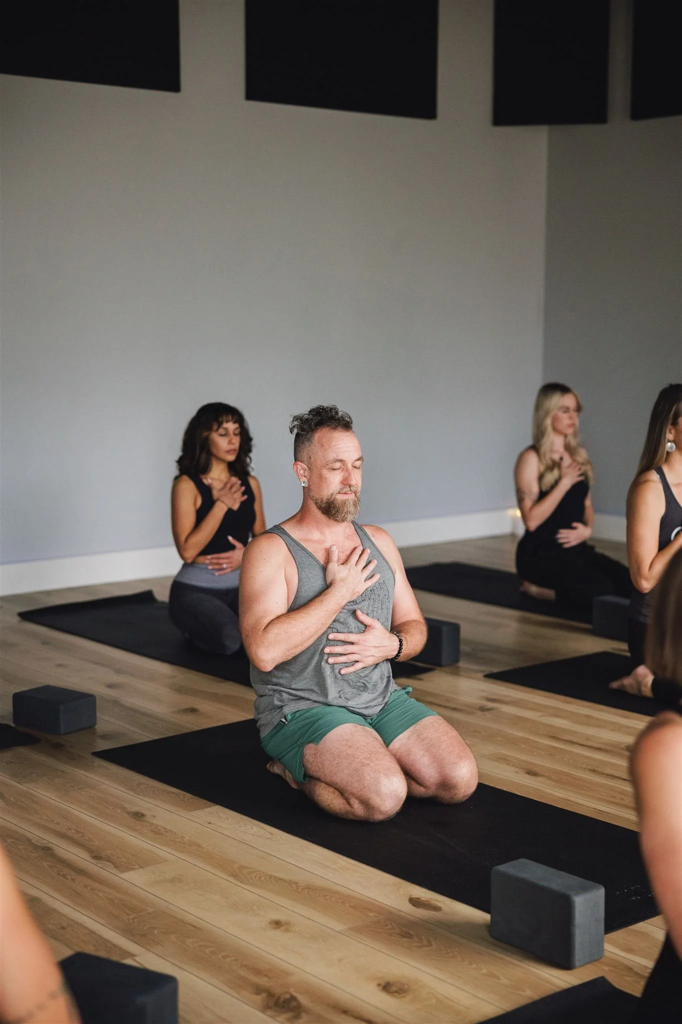 People participating in a yoga class, sitting on mats with blocks, engaging in meditation or prayer in a serene indoor studio.