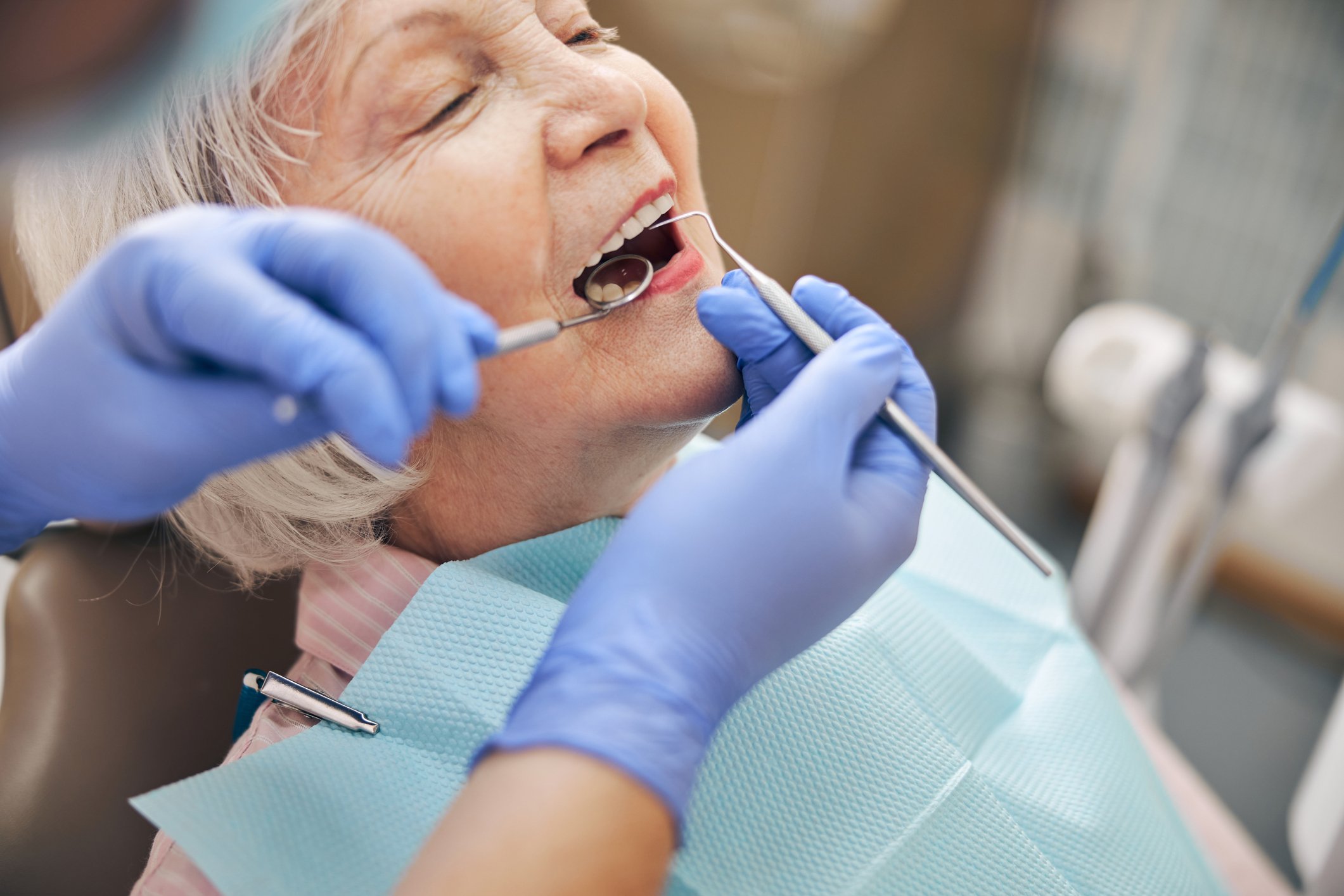 A dentist wearing blue gloves examining a smiling elderly woman patient’s open mouth with dental tools in a dental office.