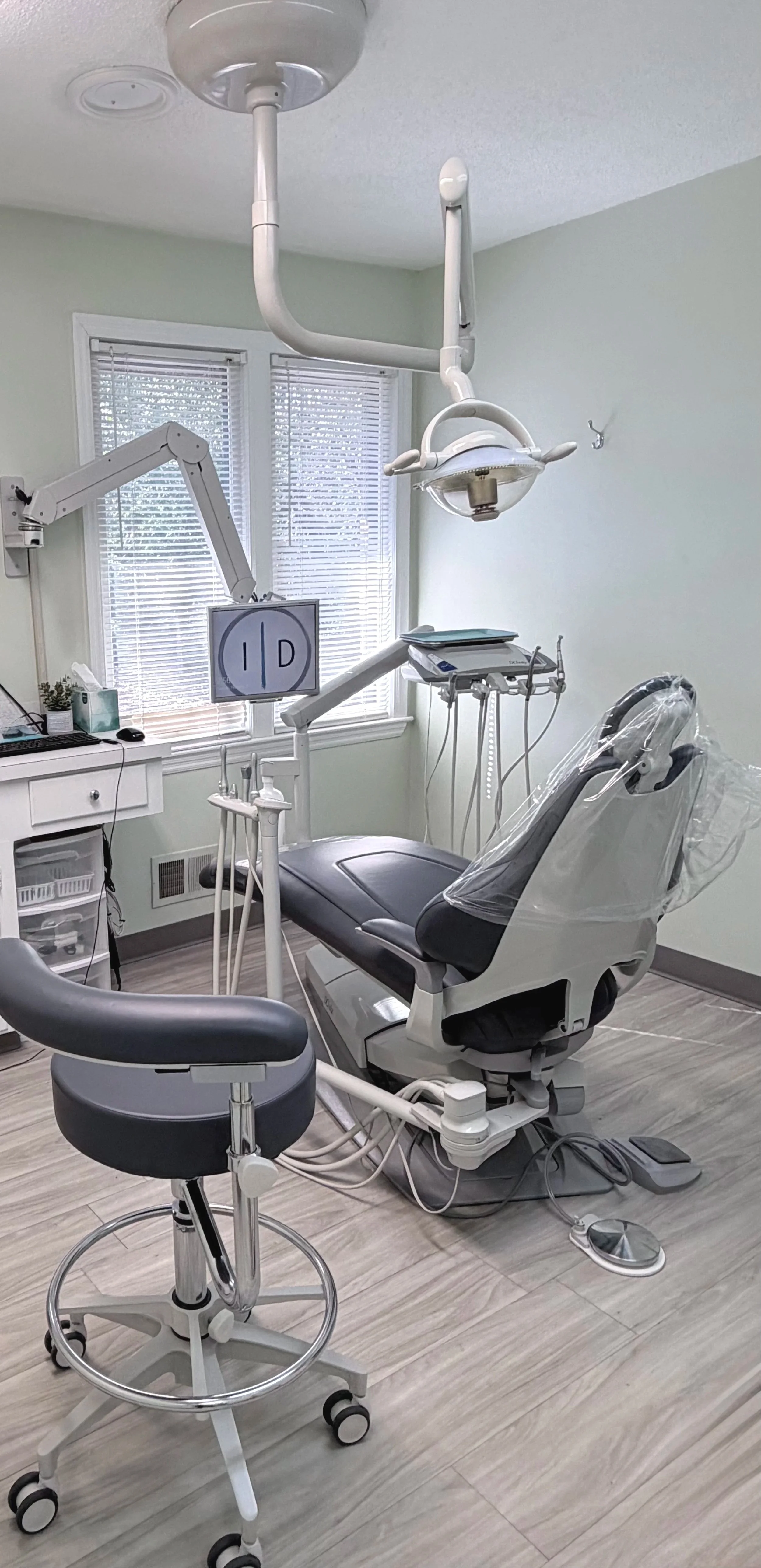 Dental examination room with dental chair, overhead light, dental tools, and a monitor, with windows in the background.