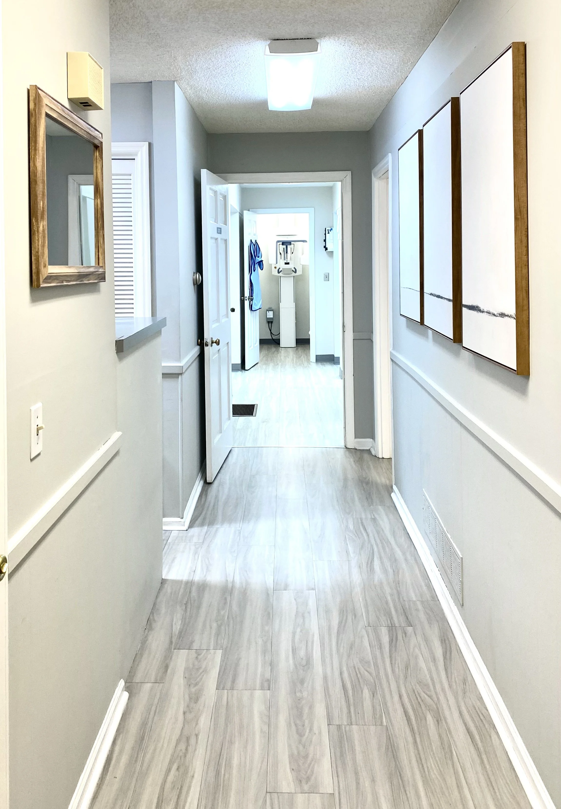 A hallway with light-colored wood flooring, white walls with trim, and artwork on the right wall. There is a mirror on the left wall and an open door leading to a room with more light and a piece of dental equipment visible in the background.