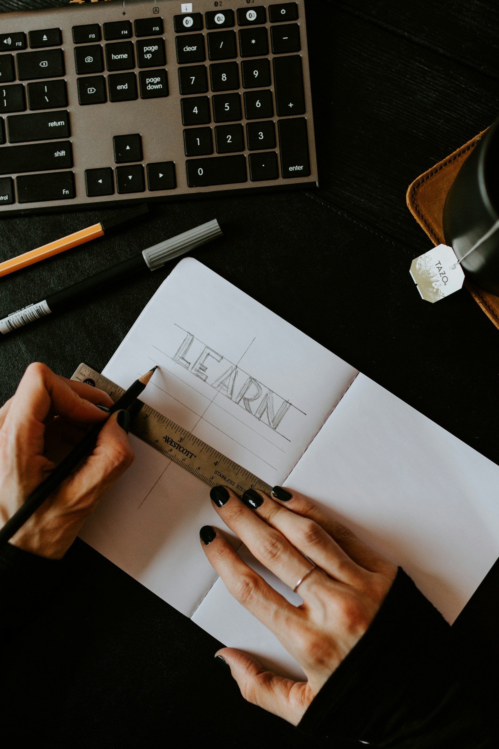 A person is sketching the word 'LEARN' in a notebook, using a ruler for alignment. The workspace includes a keyboard, pens, a tea tag, and a coffee mug.