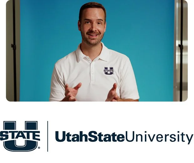 A white man with light brown hair, a mustache and beard wearing a suit and light tie smiles. at the camera in front of a yellow background above the Utah State University logo and name.