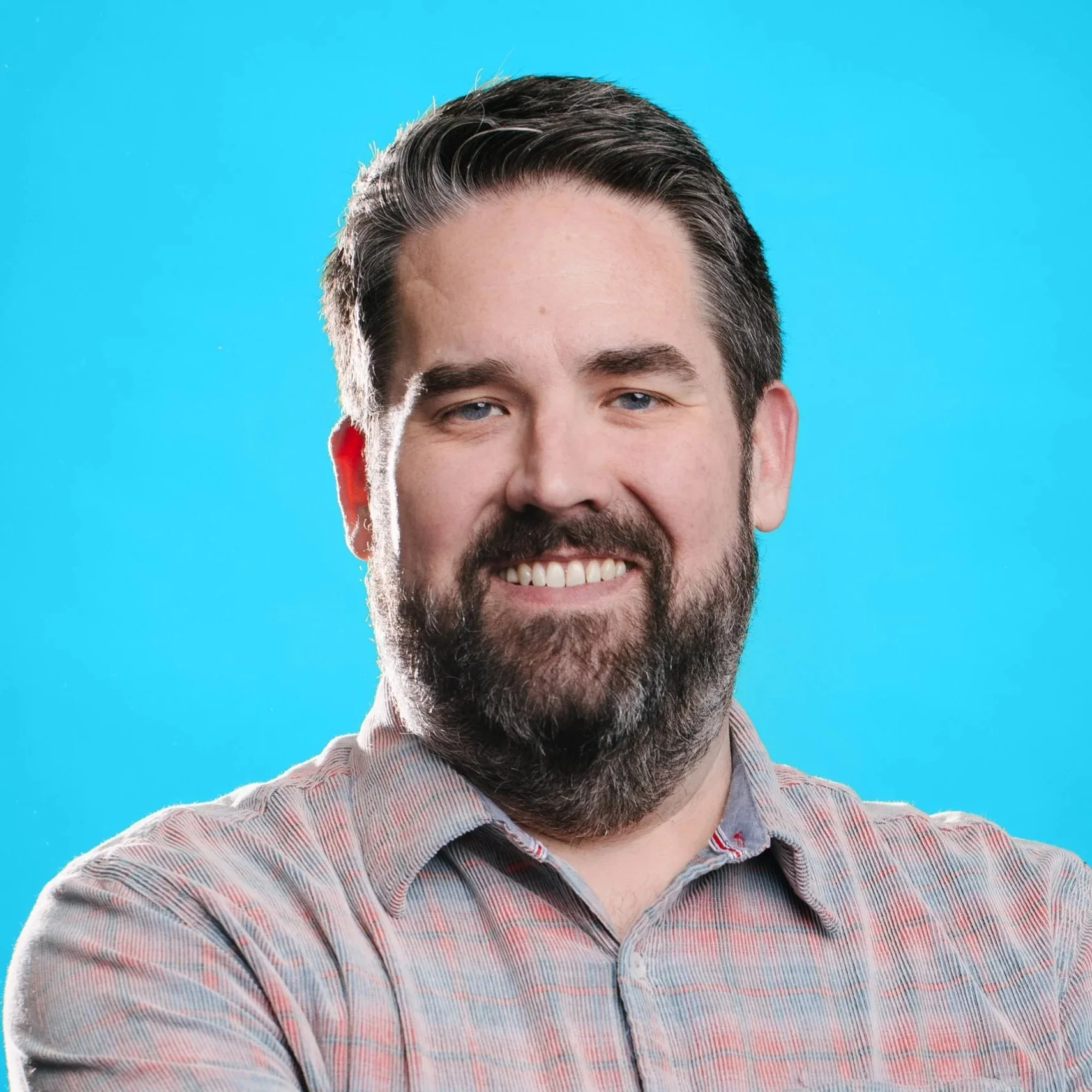 A white man with dark hair, a beard and mustache, and dark glasses smiles at teh camera. He wears a gray suit with a lavender collared shirt and a neutral background.