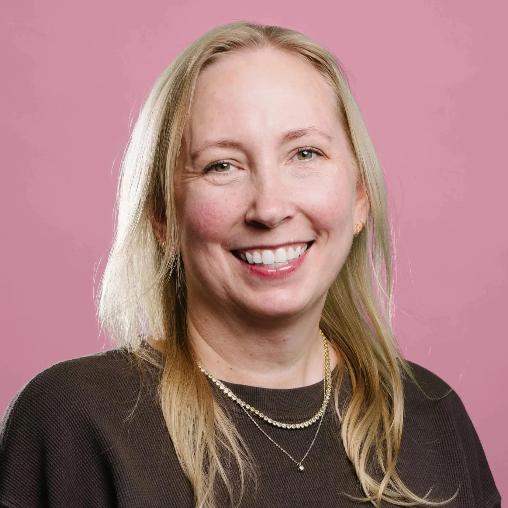 A blonde white woman smiles at the camera. She wears a light pink shirt with a wooden background behind her