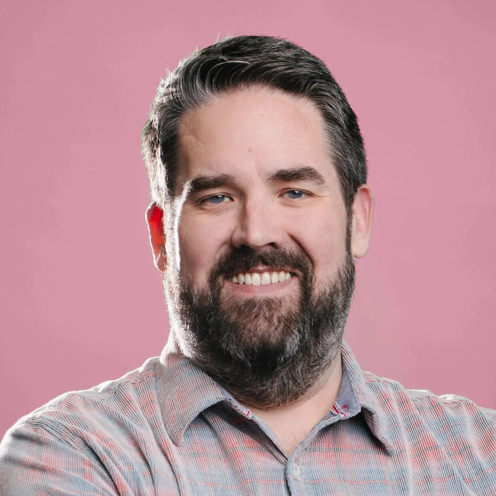 A white man with dark hair, a beard and mustache, and dark glasses smiles at teh camera. He wears a gray suit with a lavender collared shirt and a neutral background.