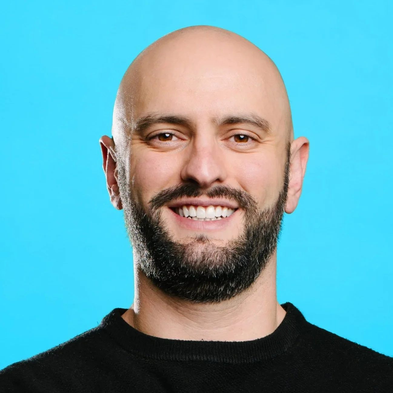 A white man with dark hair and a beard smiles directly at the camera. He wears a blue suit jacket with a white collared shirt and tie with a dark background.