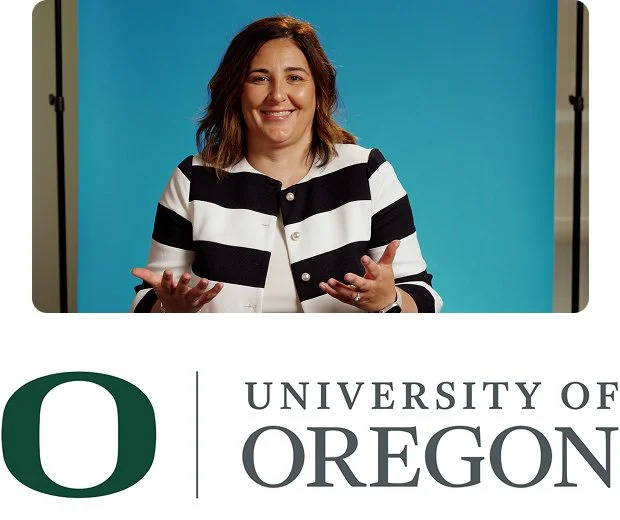 A white woman with long brown hair smiles at the camera. She wears a very light blue suit jacket and white shirt with a blue background above the University of Oregon logo and name.