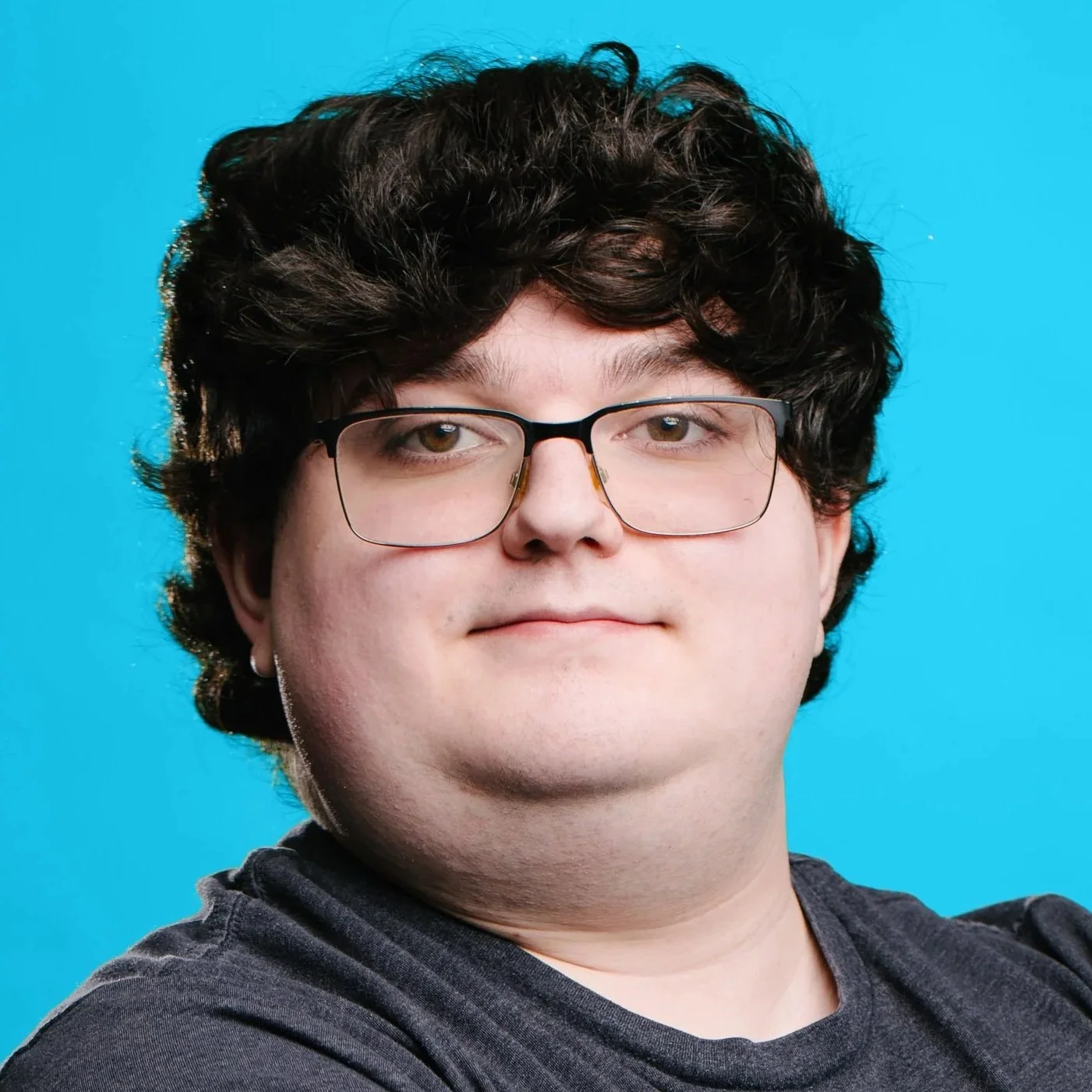 A white man with short dark hair, glasses, and light facial hair, smiling slightly and wearing a light grey t-shirt, in front of a beige wall.