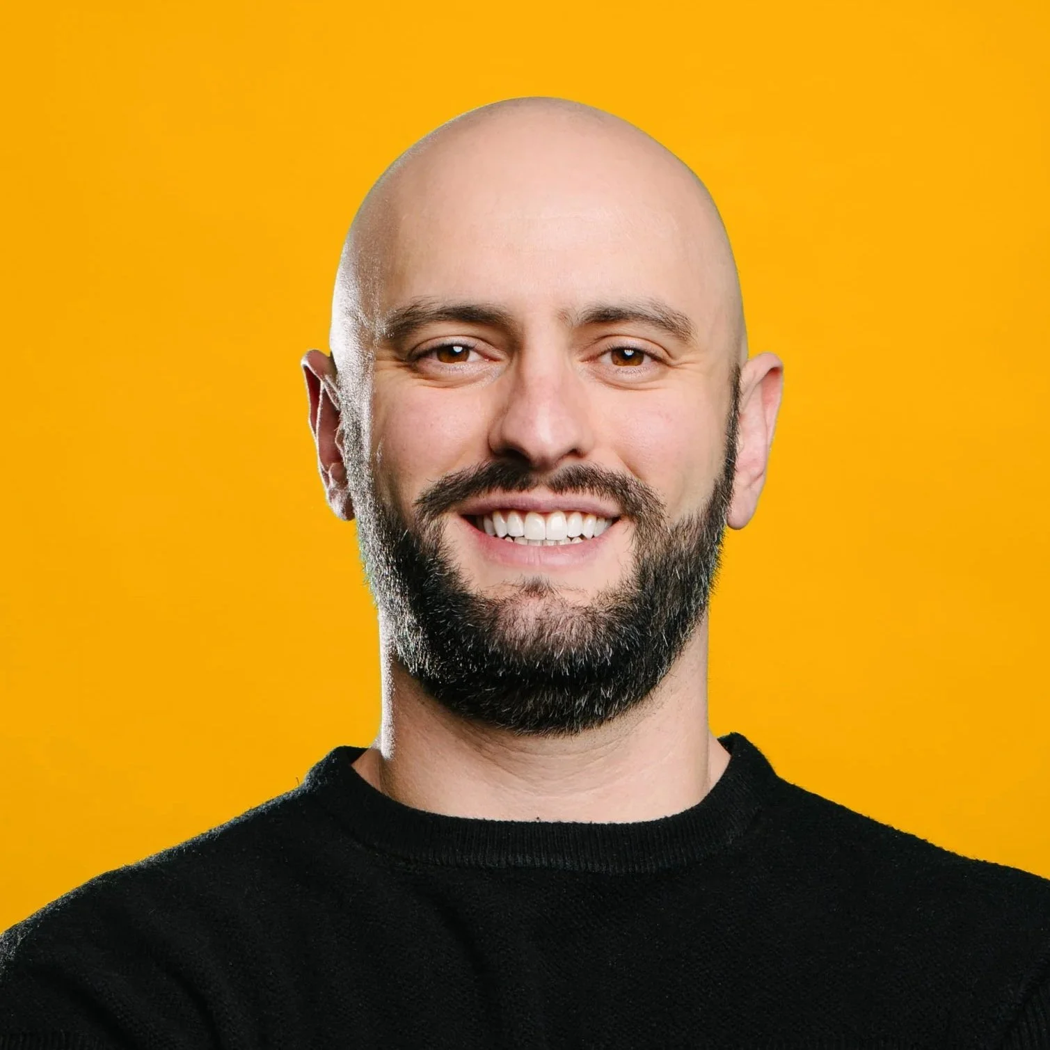 A white man with dark hair and a beard smiles directly at the camera. He wears a blue suit jacket with a white collared shirt and tie with a dark background.